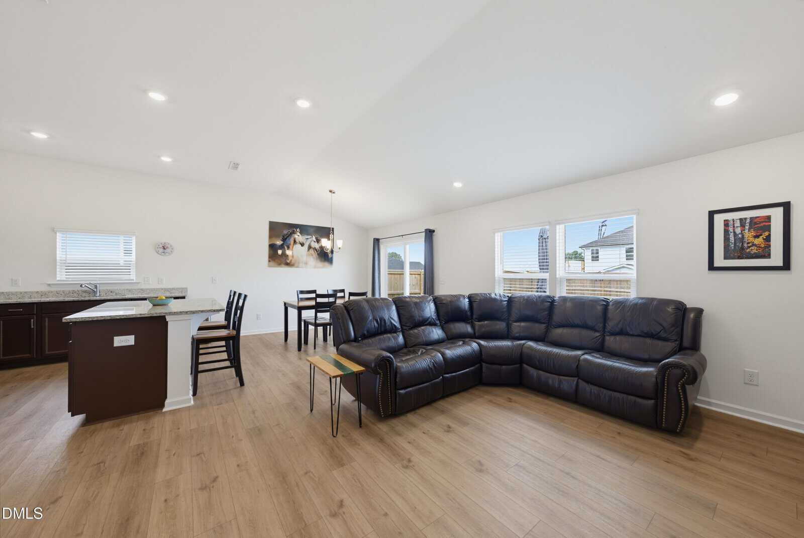 Modern open-concept living area with dark leather sectional, dining table, kitchen island, wood flooring, white walls, large windows, and a framed picture on the wall. Natural light fills the space.