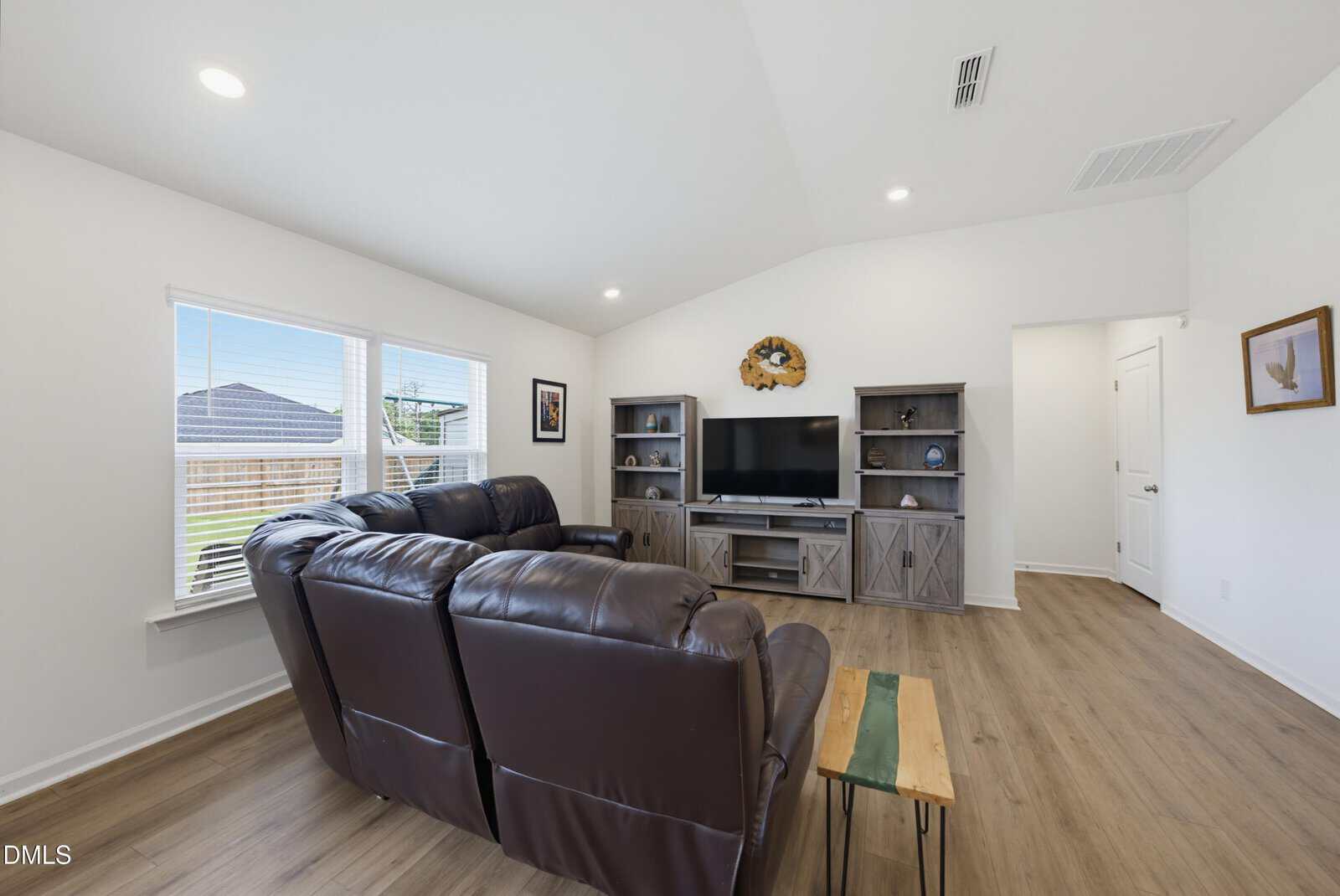 Living room with brown leather sectional sofa, wooden coffee table, and TV on a stand between two matching shelves. Large window lets in natural light, and the room has light wood floors and white walls.