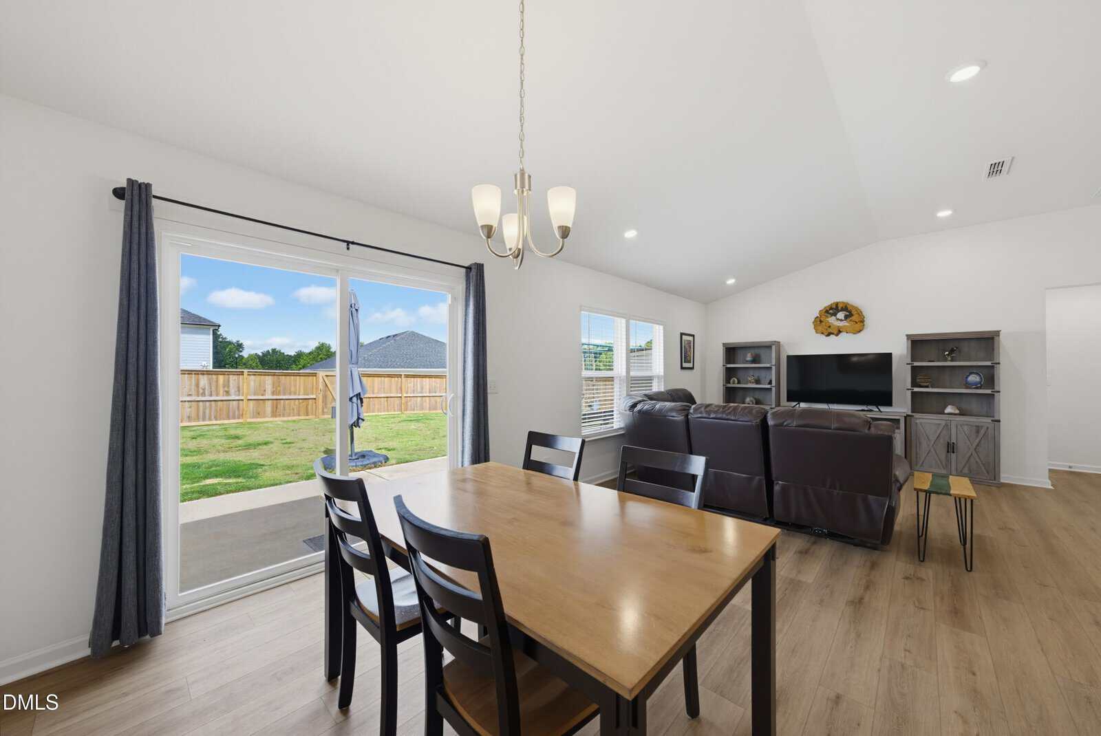 A dining area with a wooden table and four chairs is next to a living space with a brown sectional sofa facing a TV. Sliding glass doors open to a fenced backyard. The room has white walls, wood flooring, and a ceiling light.