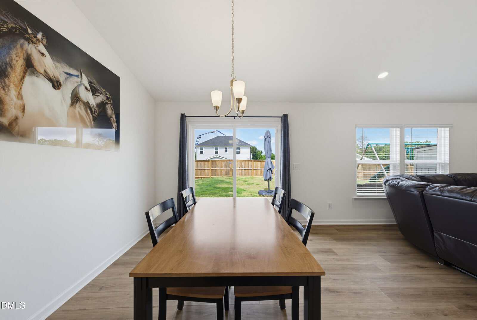 A dining area with a wooden table and four black chairs, a horse painting on the wall, chandelier above the table, sliding glass doors leading to a backyard, and a partial view of a living room sofa.