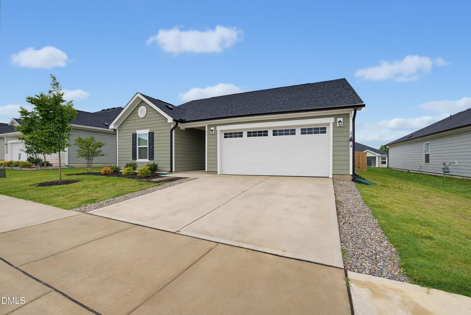 Single-story house with light green siding, dark roof, white trim, and double garage. Concrete driveway, small front lawn with landscaping, and neighboring homes visible under a blue sky with scattered clouds.