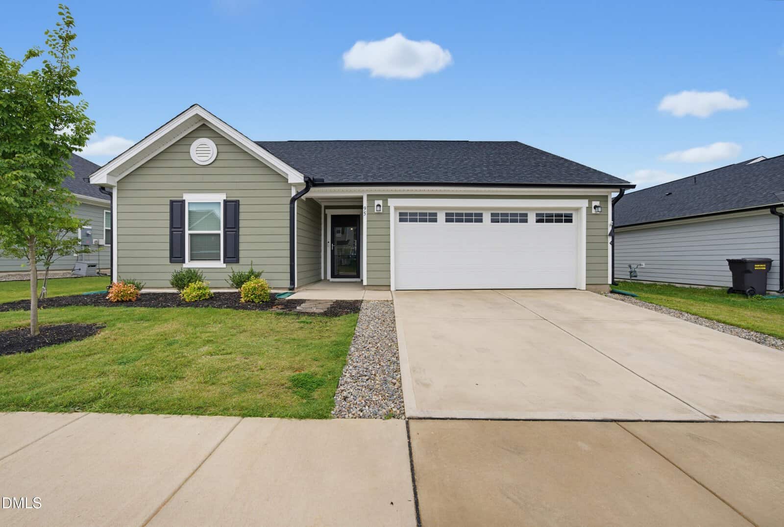 A single-story house with light green siding, white trim, and a two-car garage. There is a concrete driveway, small landscaped front yard with shrubs, and a tree on the left. The sky is blue with a few clouds.