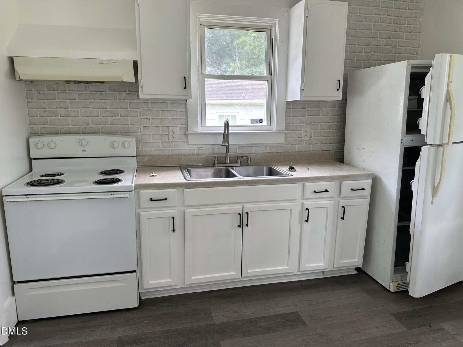 A small kitchen with white cabinets, an electric stove, a double sink, and an open refrigerator. There is a window above the sink and gray wood flooring.