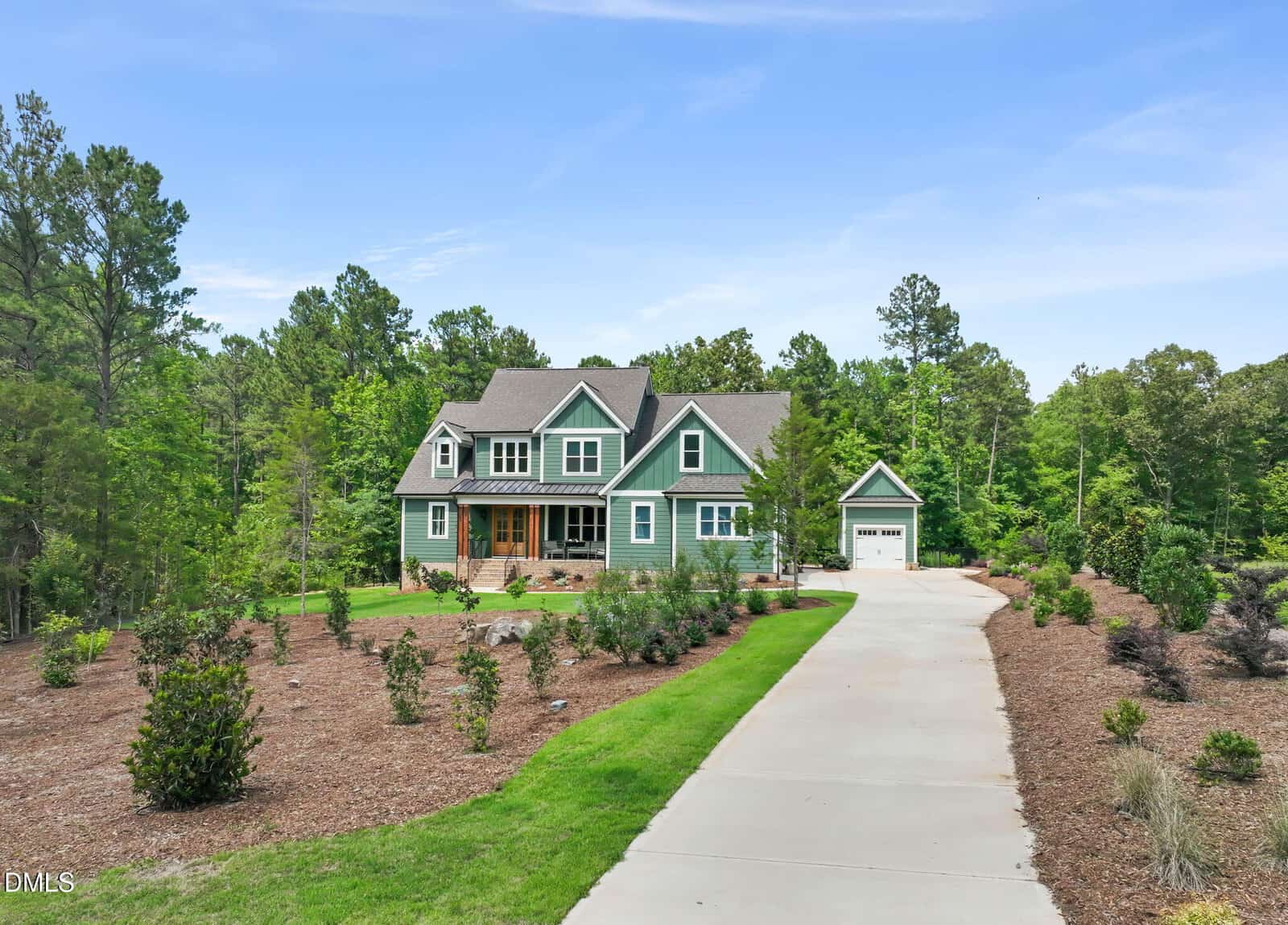 Nestled in Louisburg, 270 Whistlers Cove is a two-story green house with white trim and a covered front porch, set amid landscaped gardens and trees. A long driveway leads to a detached garage on the right under a clear blue sky.