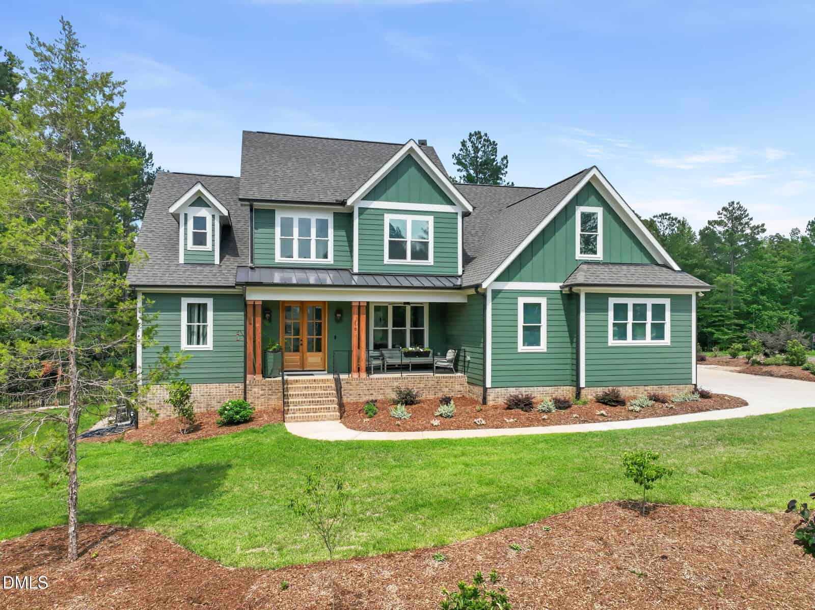 Two-story green house with white trim, a covered front porch, wooden double doors, black roof, and a curved driveway, surrounded by green lawn, mulch, and trees under a blue sky.