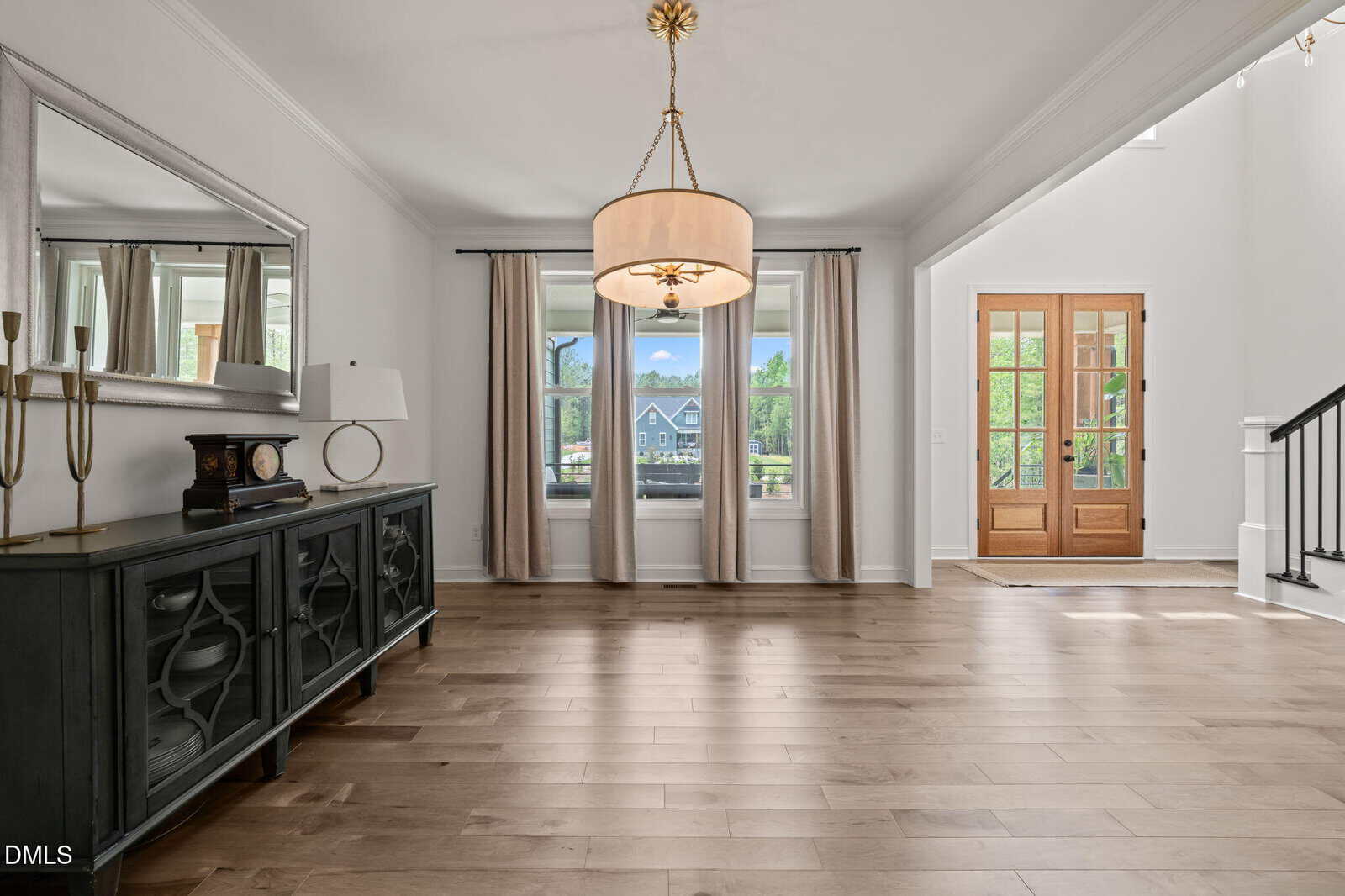 Bright entryway with light wood floors, a black sideboard and mirror on the left, a large round chandelier, and double wooden front doors with glass panels. Big window shows a house and greenery outside.