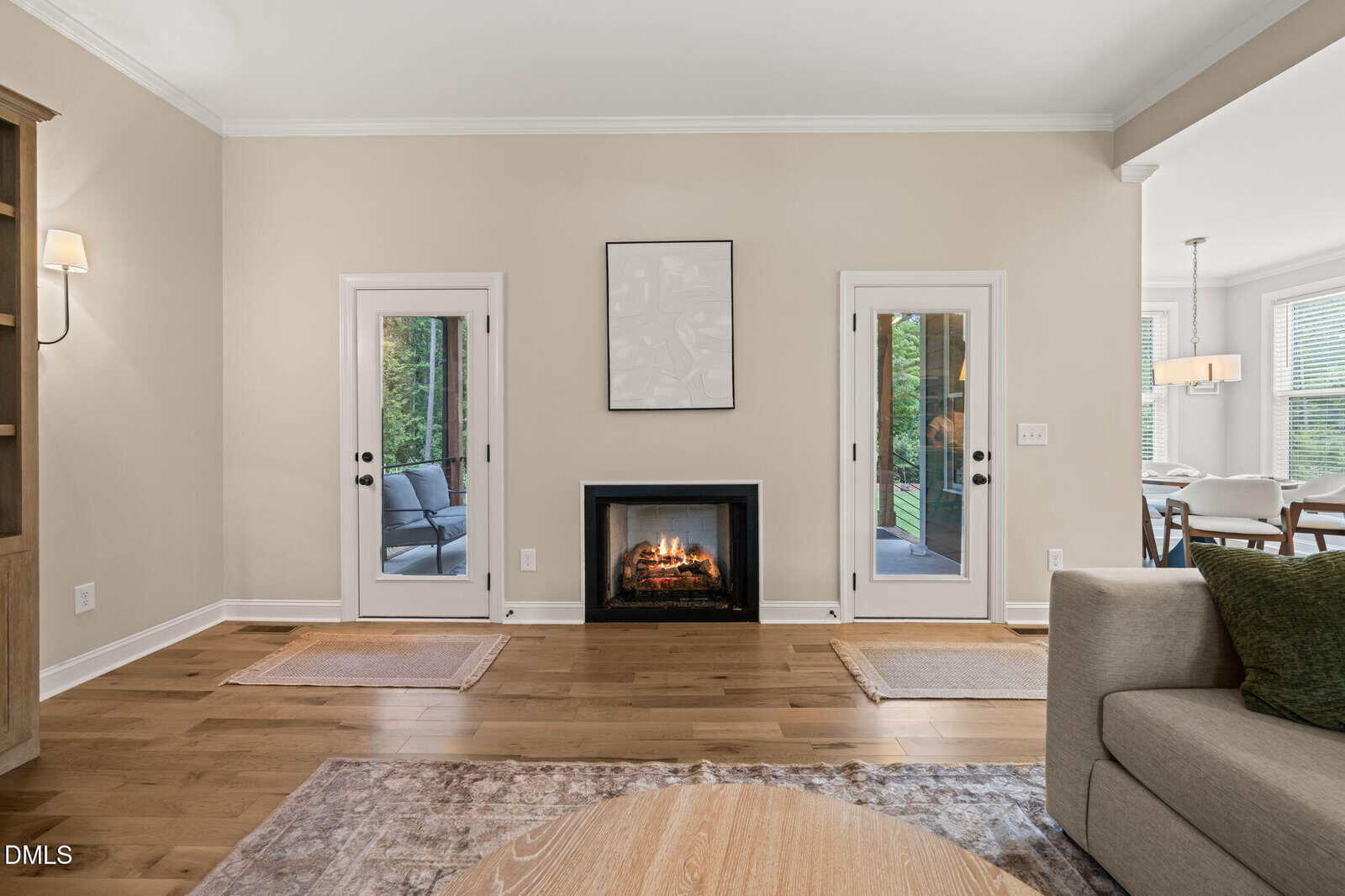 Living room with a fireplace centered between two glass doors, light beige walls, wood flooring, and a partial view of a dining area on the right. A neutral artwork hangs above the fireplace.
