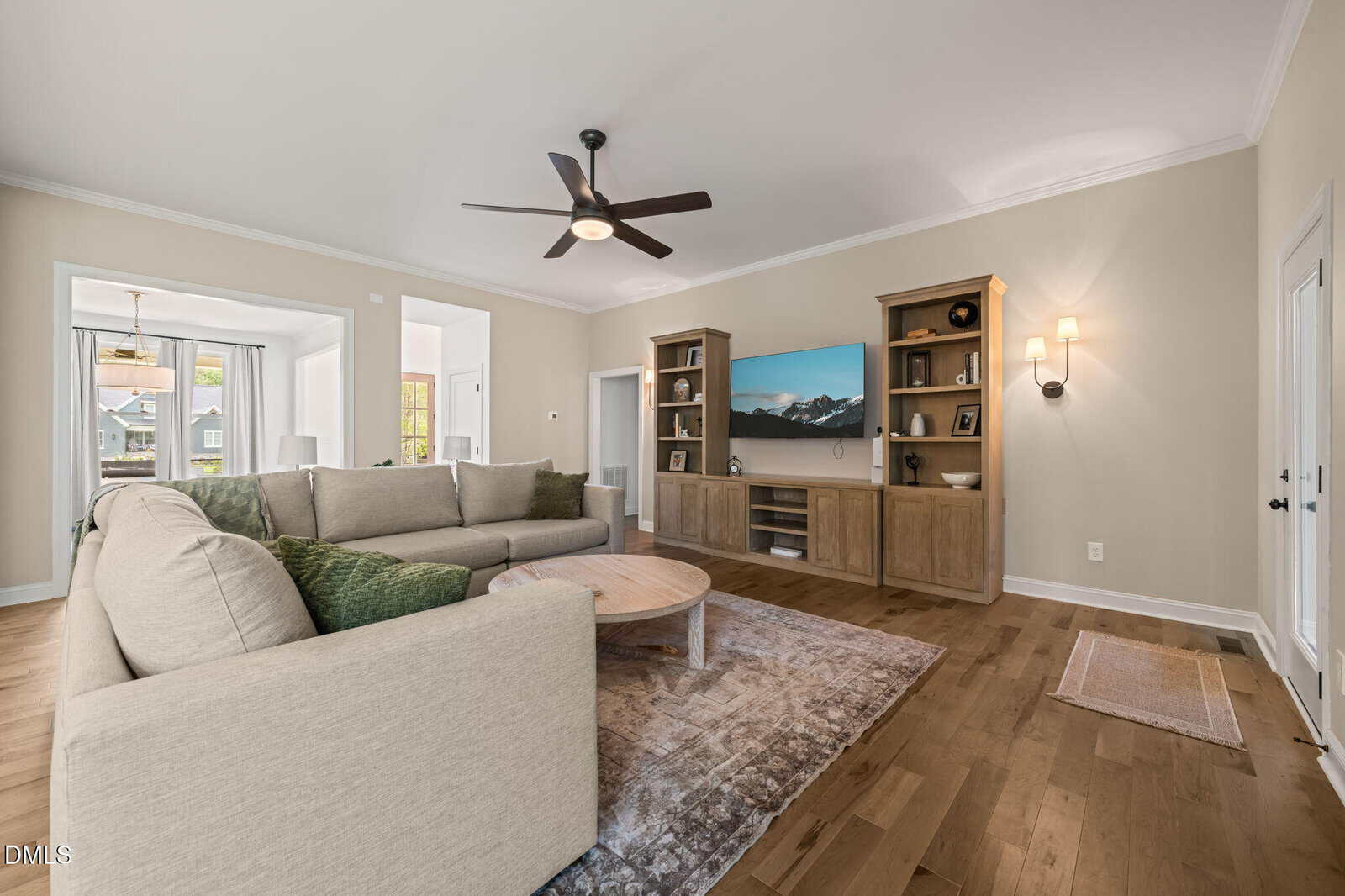 Modern living room with beige sectional sofa, wooden floors, a round coffee table, built-in shelves with a TV, wall sconces, and large windows letting in natural light. Neutral color palette with green accent pillows.