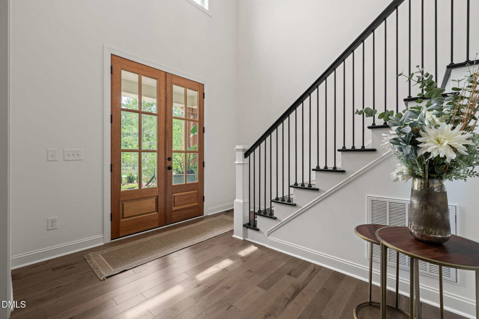 A bright entryway features double wooden front doors with glass panels, a staircase with black railings, light wood flooring, a doormat, and a small round table with a floral arrangement.