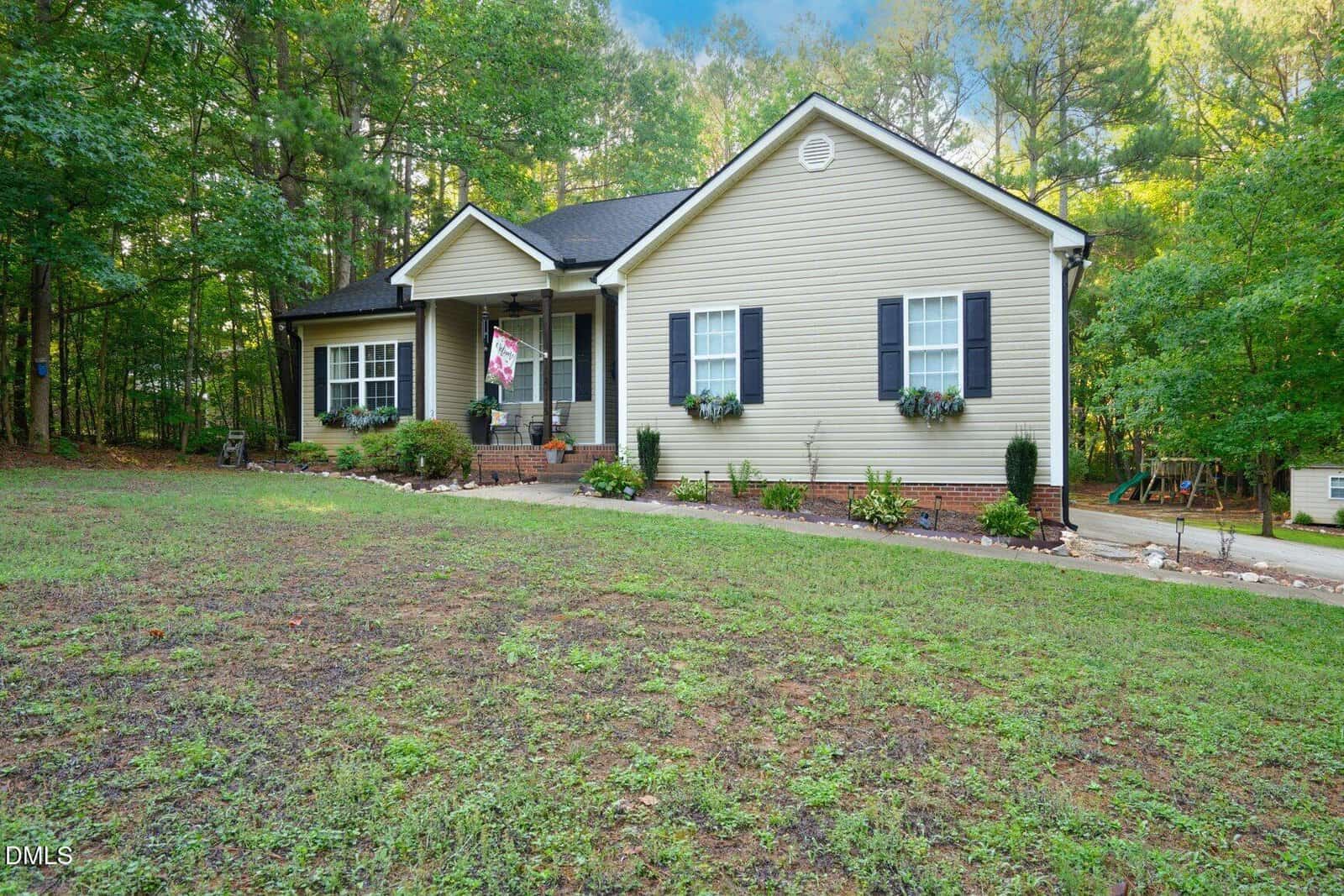 Charming single-story beige house at 4115 Geneva Drive, Franklinton, with black shutters, a covered front porch, and flower boxes. Surrounded by trees and a large grassy yard with a driveway along the right side.