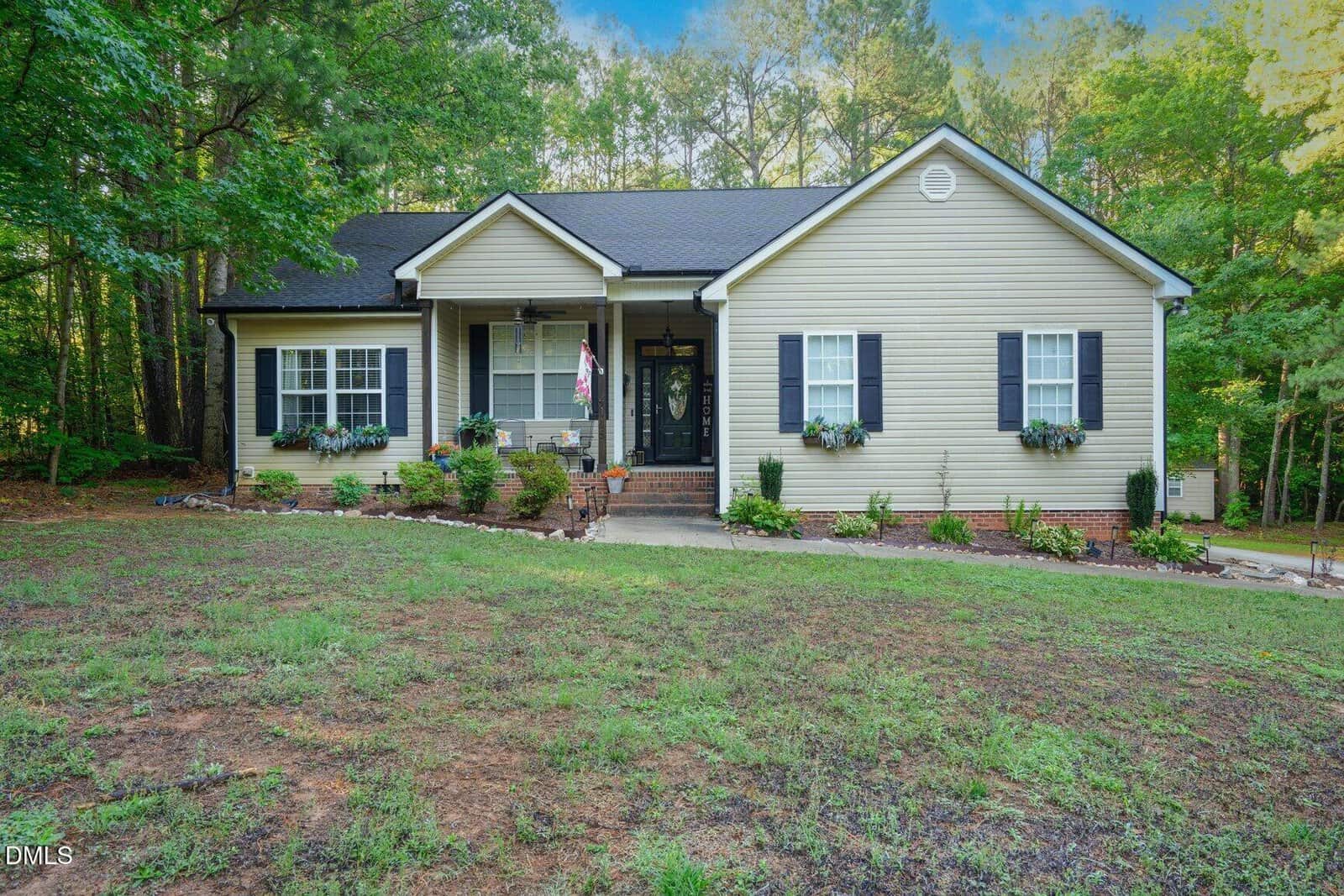 Single-story beige house with black shutters and flower boxes, set on a slightly sloped yard with patchy grass, surrounded by tall green trees under a blue sky.
