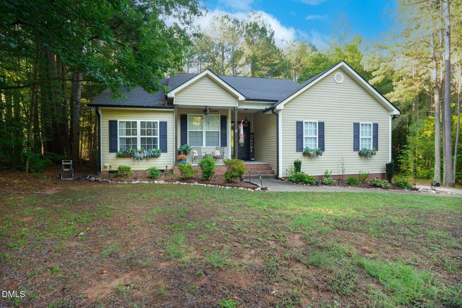 Single-story beige house with dark shutters and white trim, surrounded by trees. A small front porch with potted plants, flower boxes under windows, and a neatly landscaped yard with grass and mulch beds.