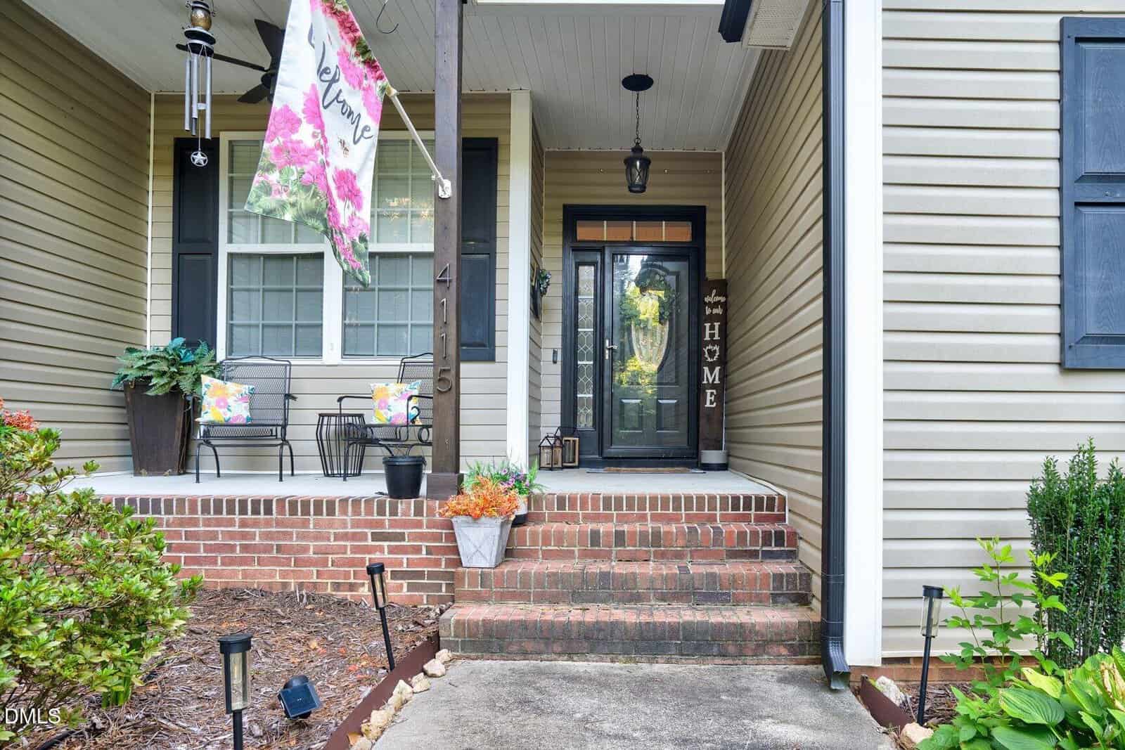 Front porch with brick steps, a glass door, potted plants, welcome flag, two chairs with floral cushions, and a “HOME” sign leaning by the door. Tan siding and black shutters are visible.