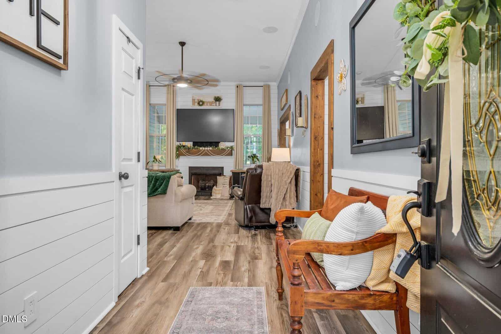 A cozy entryway leads to a living room with a wooden bench, pillows, and a wreath on the door. The living room features a sofa, armchairs, a fireplace, and a TV, with large windows letting in natural light.