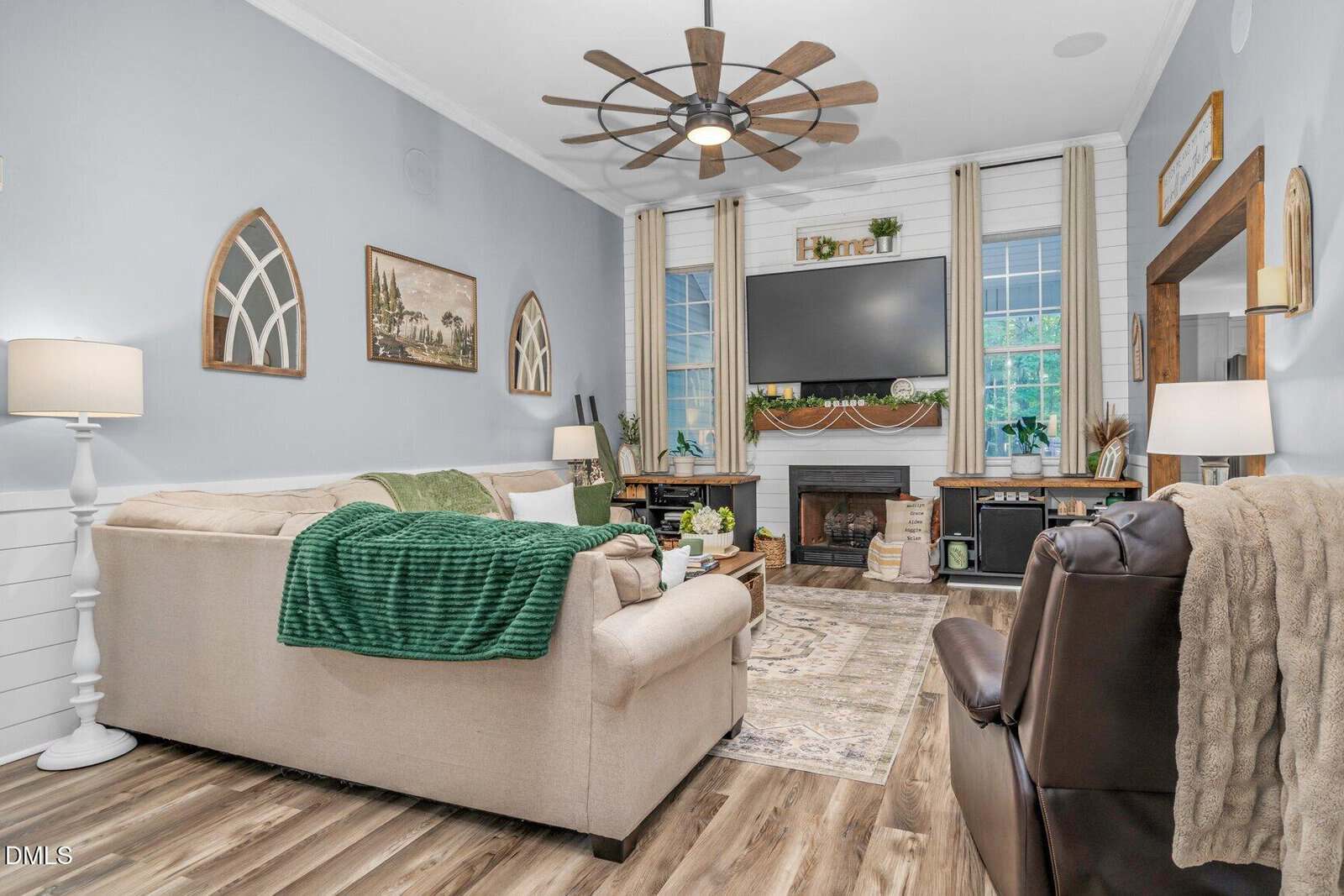 Cozy living room with neutral sofas, a green throw, large ceiling fan, TV above a fireplace, wood floors, large windows, and farmhouse decor with plants and a “Home” sign on the mantle.