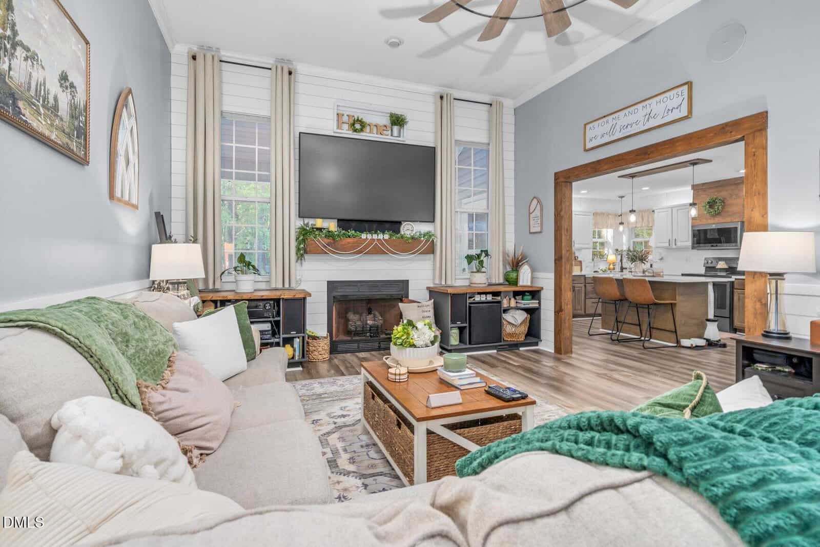 Bright, cozy living room with a beige sectional sofa, green throw blankets, a wall-mounted TV above a fireplace, and wood accents. Open doorway leads to a modern kitchen with island seating.