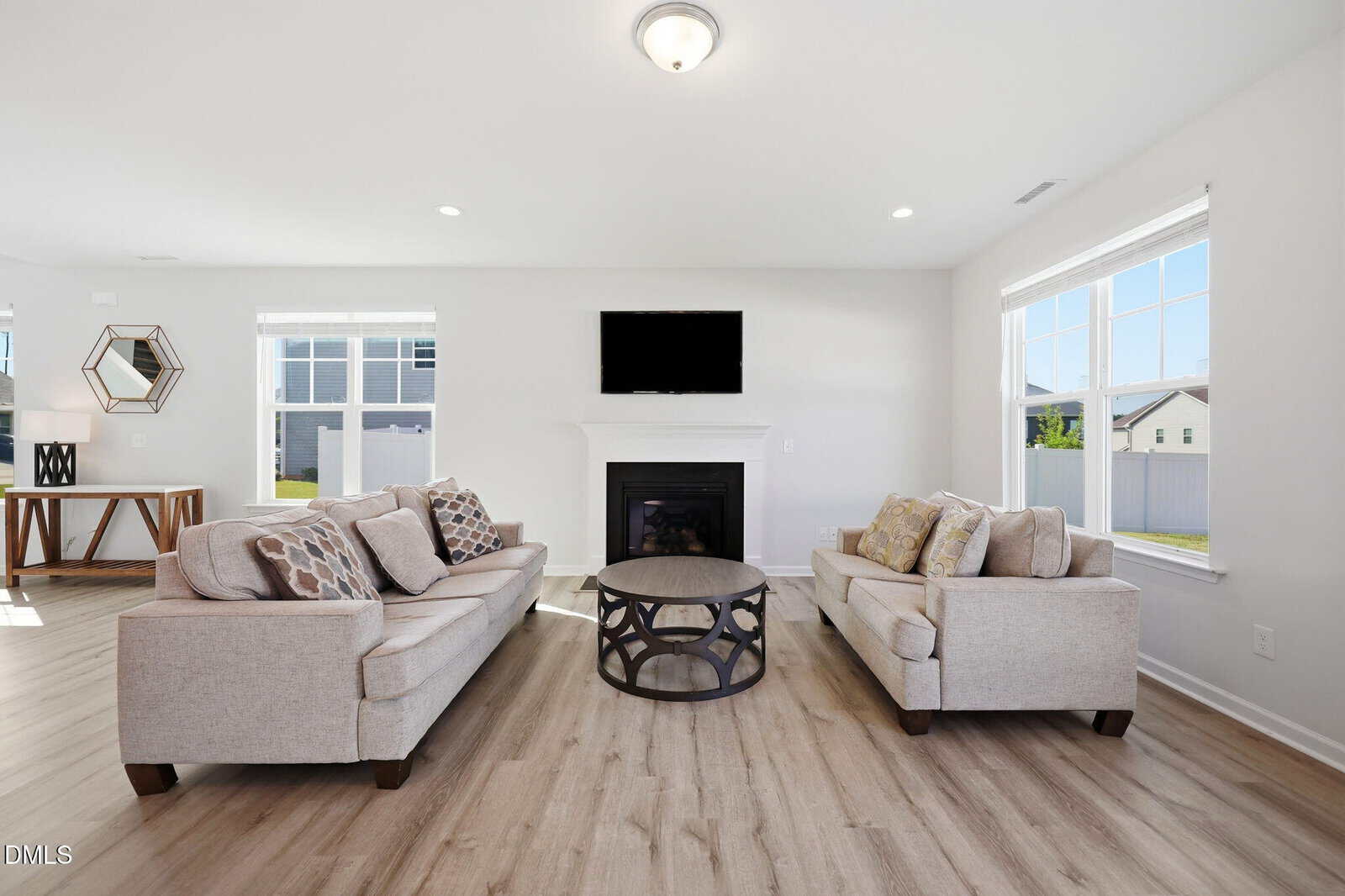 Bright living room with two beige sofas facing a round coffee table and a fireplace with a wall-mounted TV. Large windows let in natural light, and wood flooring adds warmth to the space.