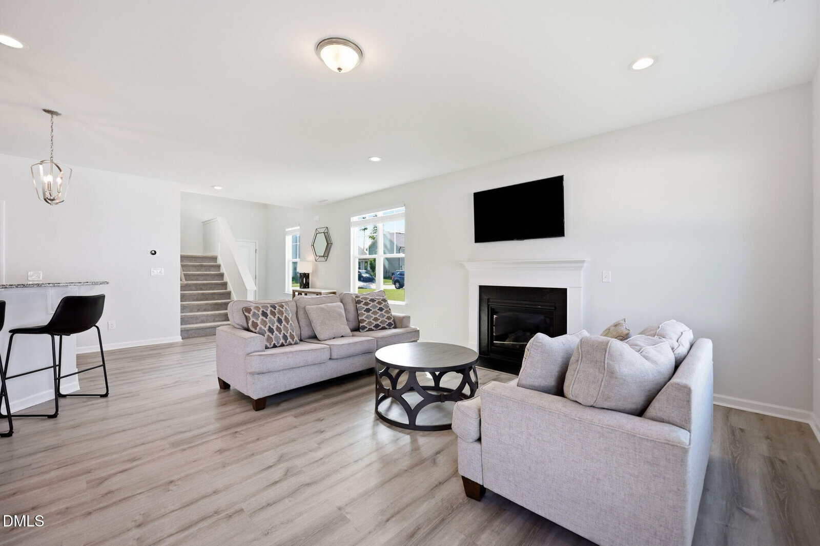 Bright, modern living room with two gray sofas, a round coffee table, a wall-mounted TV above a fireplace, and an open view of stairs and kitchen area with bar stools. Large windows provide natural light.