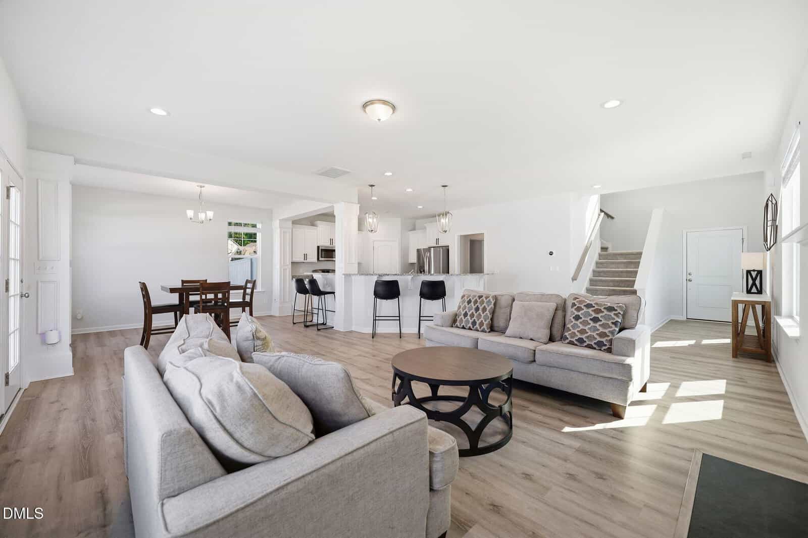 Bright, open-concept living space with light wood floors, two gray sofas, a round coffee table, a dining area, modern kitchen with bar stools, and a staircase leading upstairs. Large windows let in natural light.