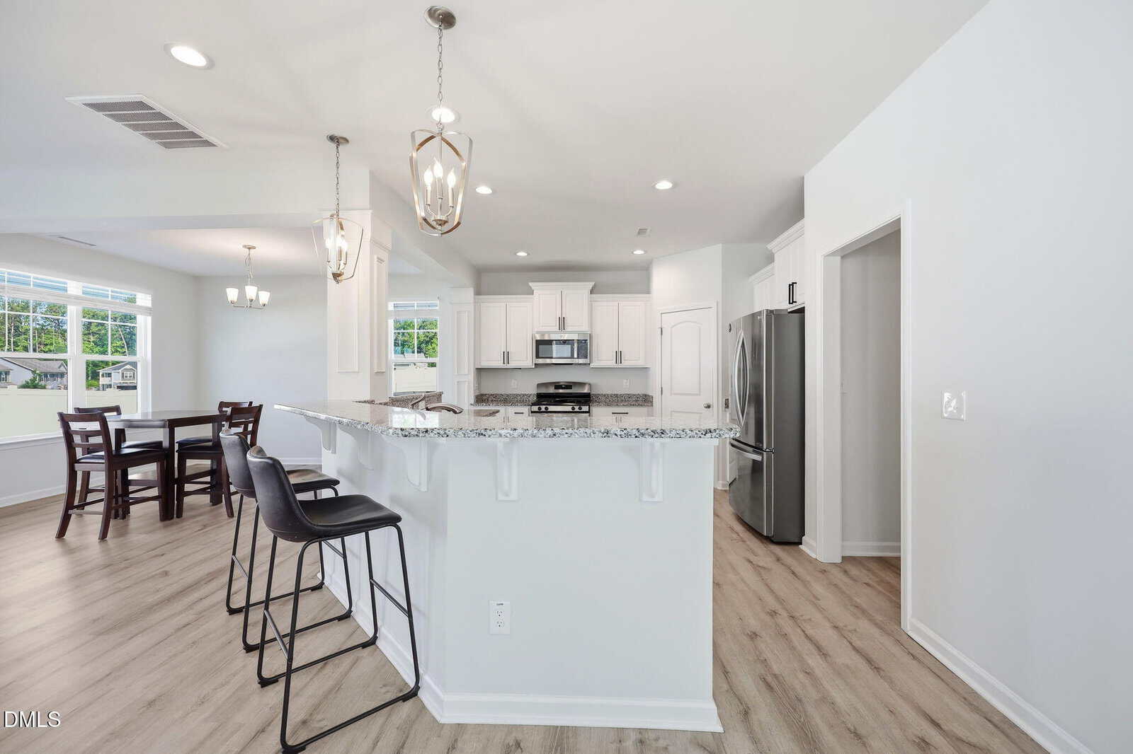 Bright, modern kitchen with white cabinets, stainless steel appliances, granite countertops, light wood floors, two pendant lights over a breakfast bar with two black stools, and an open dining area with a table and chairs.
