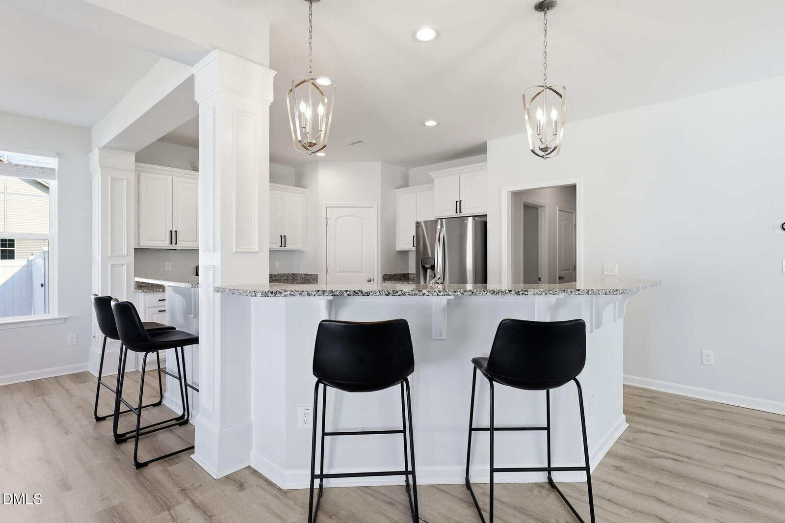 Modern white kitchen with granite countertops, stainless steel appliances, two pendant lights, and three black barstools at a breakfast bar. Light wood flooring and ample natural light from a nearby window.