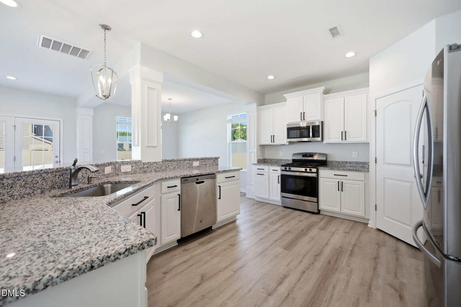 Modern kitchen with white cabinets, stainless steel appliances, granite countertops, light wood flooring, and pendant lighting. There’s a large sink, dishwasher, and ample natural light from windows in the open layout.