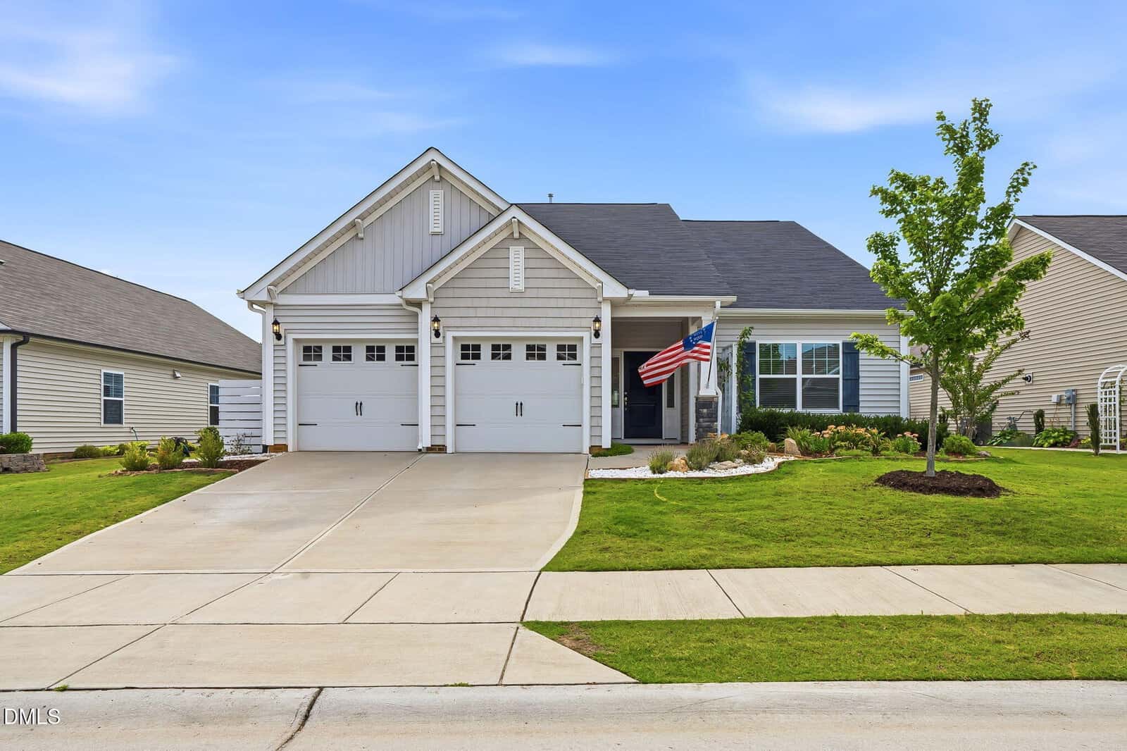 Single-story suburban house with two white garage doors, gray siding, manicured lawn, small tree, and an American flag by the front porch, under a clear blue sky.