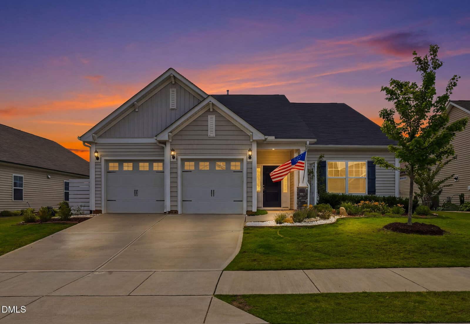 Single-story suburban house with a two-car garage, lit front porch, and an American flag, surrounded by a well-kept lawn and landscaping at sunset.