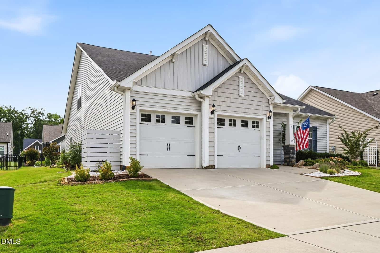 A modern suburban house with a double garage, gray siding, neatly trimmed lawn, and landscaping. An American flag is displayed to the right of the garage. The sky is clear and blue.