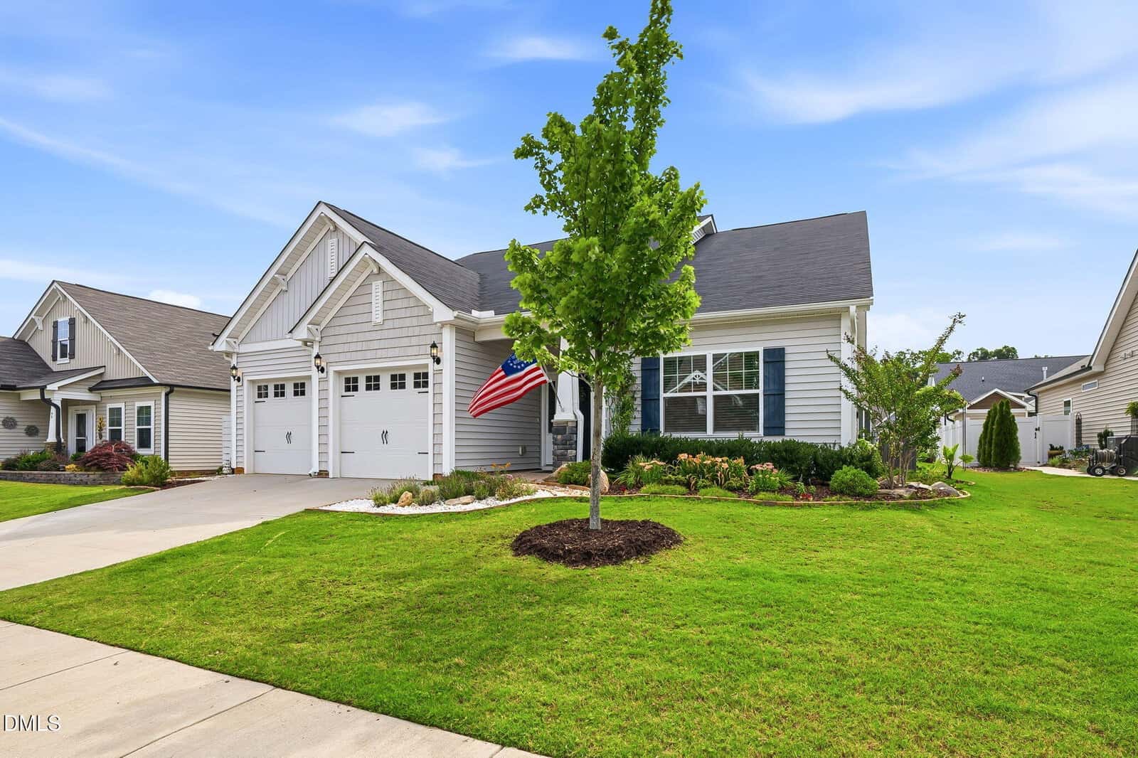 A single-story house with gray siding, white trim, and a two-car garage. An American flag hangs by the front porch. The yard is well-maintained with green grass, a tree, shrubs, and a wide driveway under a blue sky.