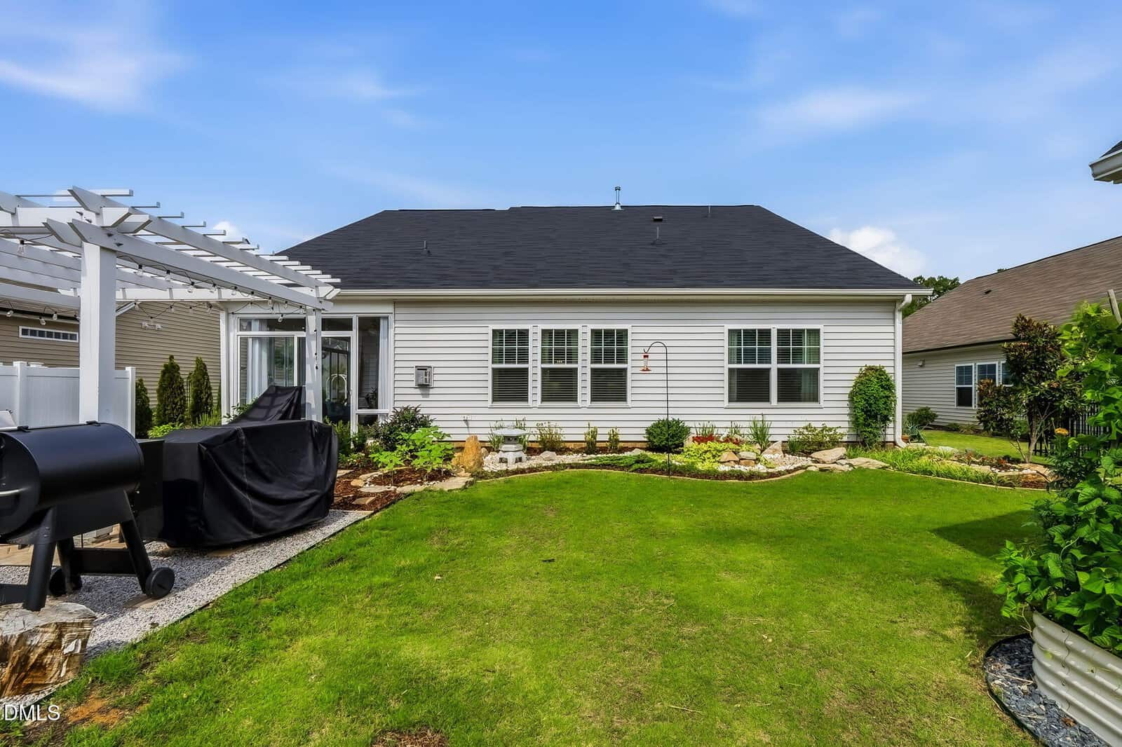 Backyard with a green lawn, a small garden, a white pergola, covered grill, and a white single-story house with large windows and a black roof under a blue sky.