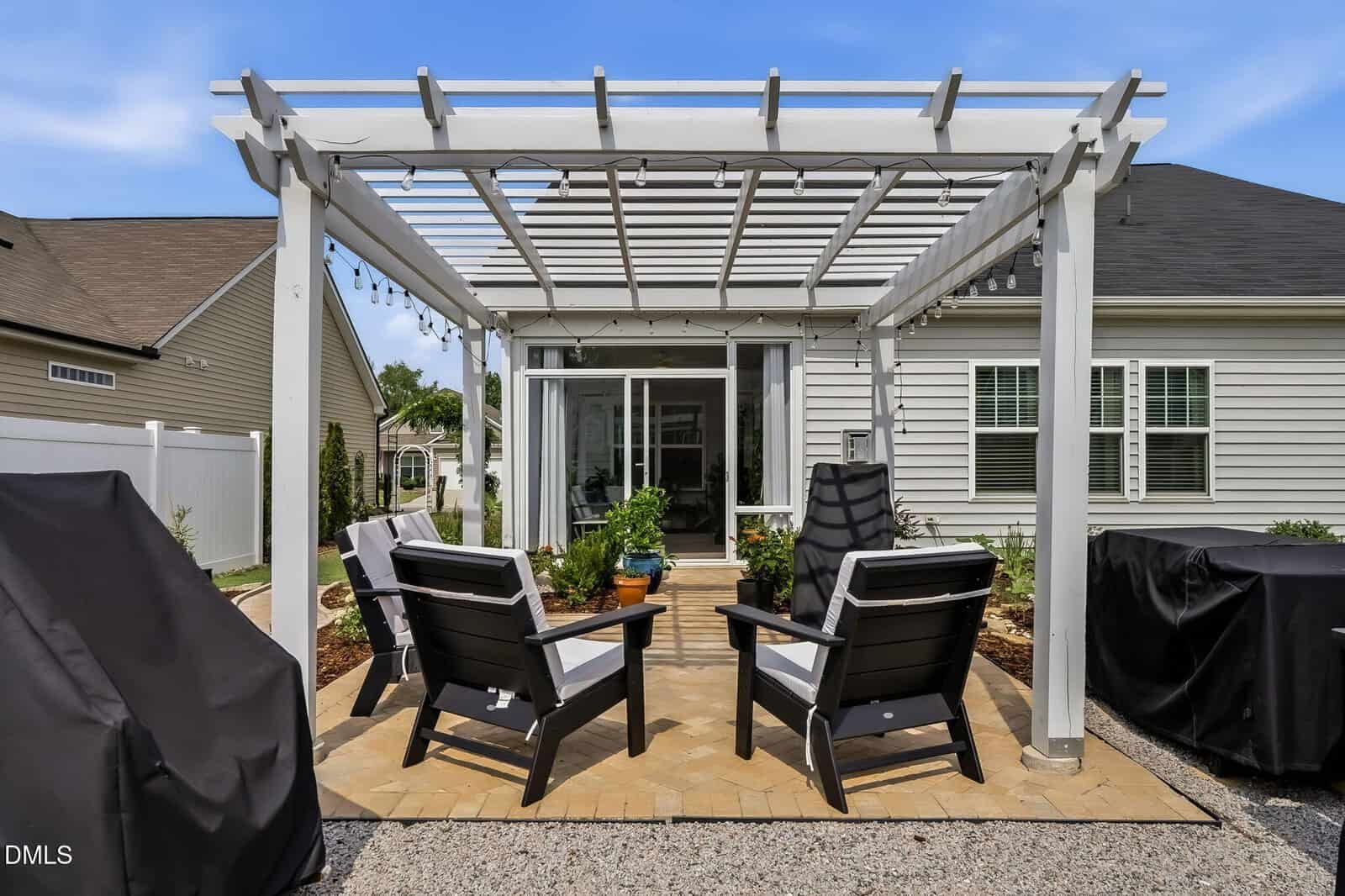 A backyard patio with a pergola, four black and white chairs, potted plants, string lights, and two covered grills. The patio sits behind a light-colored house with sliding glass doors and windows.