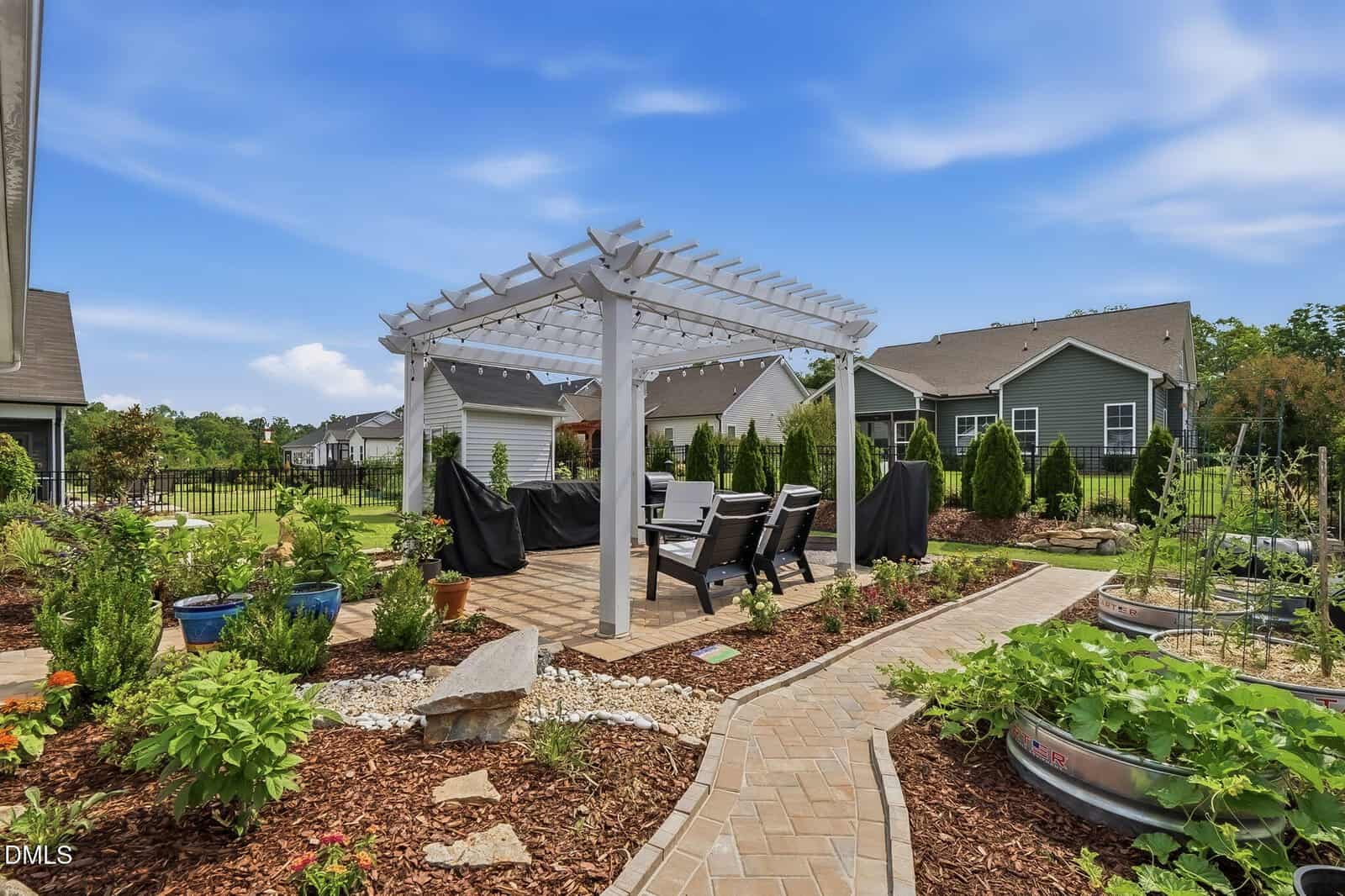A backyard patio with a white pergola, outdoor dining set, garden beds, potted plants, and a curving brick walkway under a blue sky.