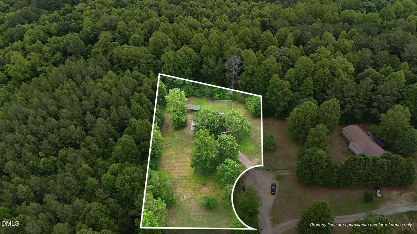 Aerial view of a wooded area with a white outline marking a section containing a house, yard, and outbuilding. Surrounding the outlined property are dense green trees and a driveway with parked vehicles.