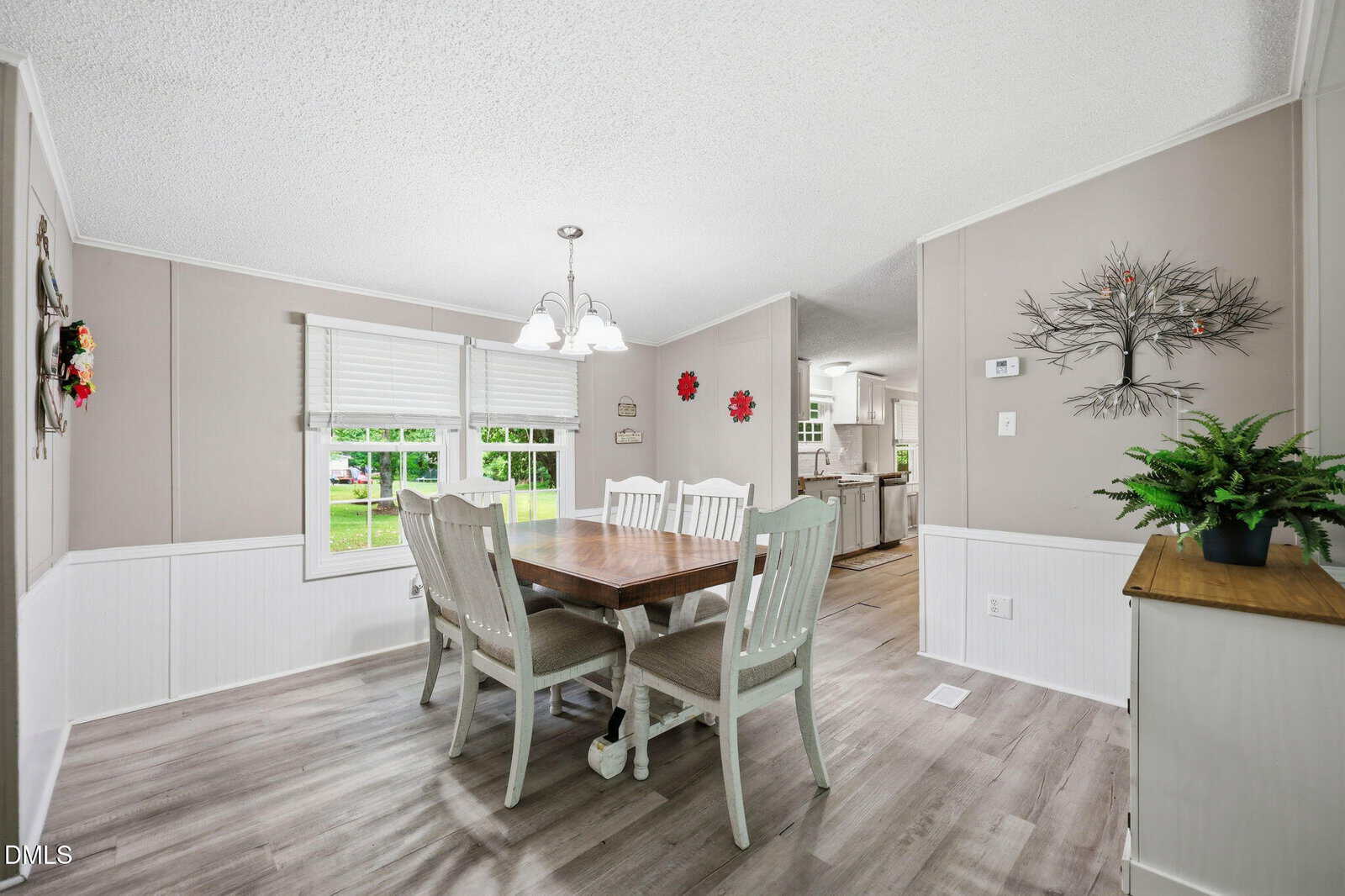 A bright dining room with a wooden table, six white chairs, light gray walls, two large windows, wall art, a plant on a sideboard, and an open view into a white kitchen.