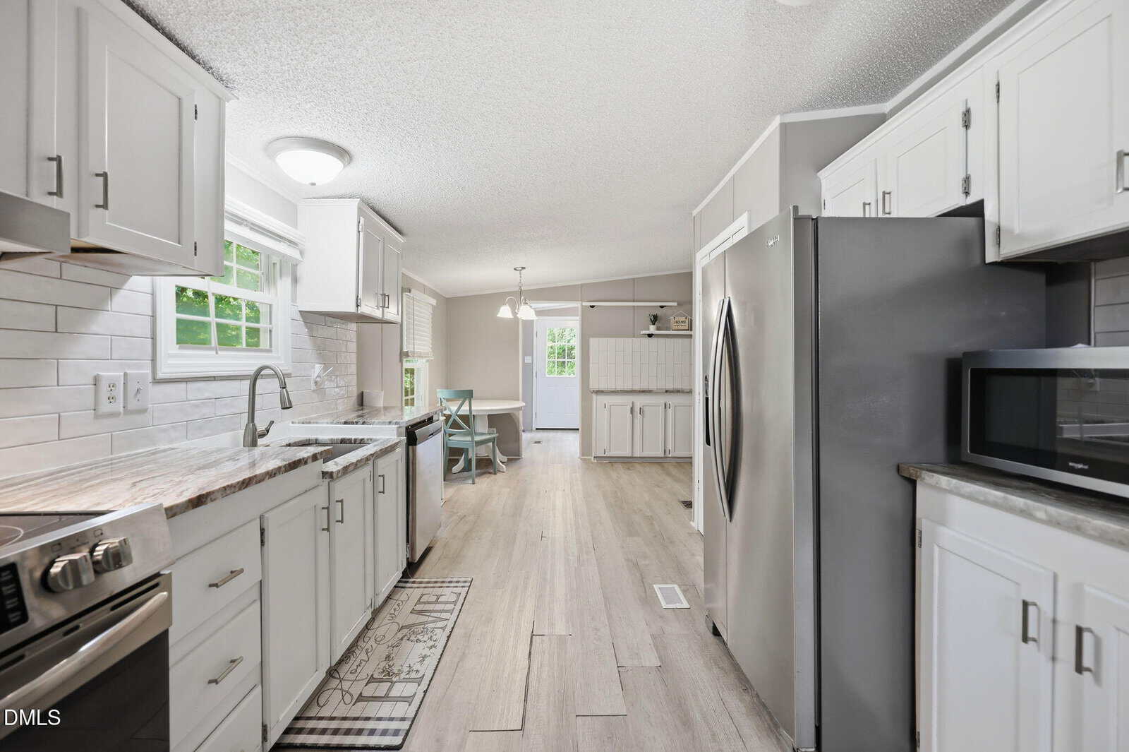 Modern kitchen with white cabinets, stainless steel appliances, a double sink under a window, light wood flooring, and a dining area with a table and chairs visible in the background.