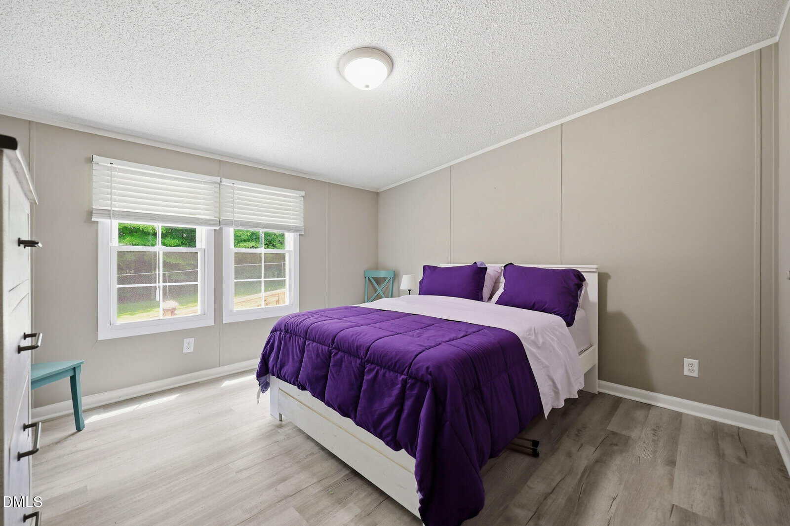 A simple bedroom with a bed featuring purple bedding, two windows with white blinds, light gray walls, wood flooring, and a turquoise side chair near the corner.