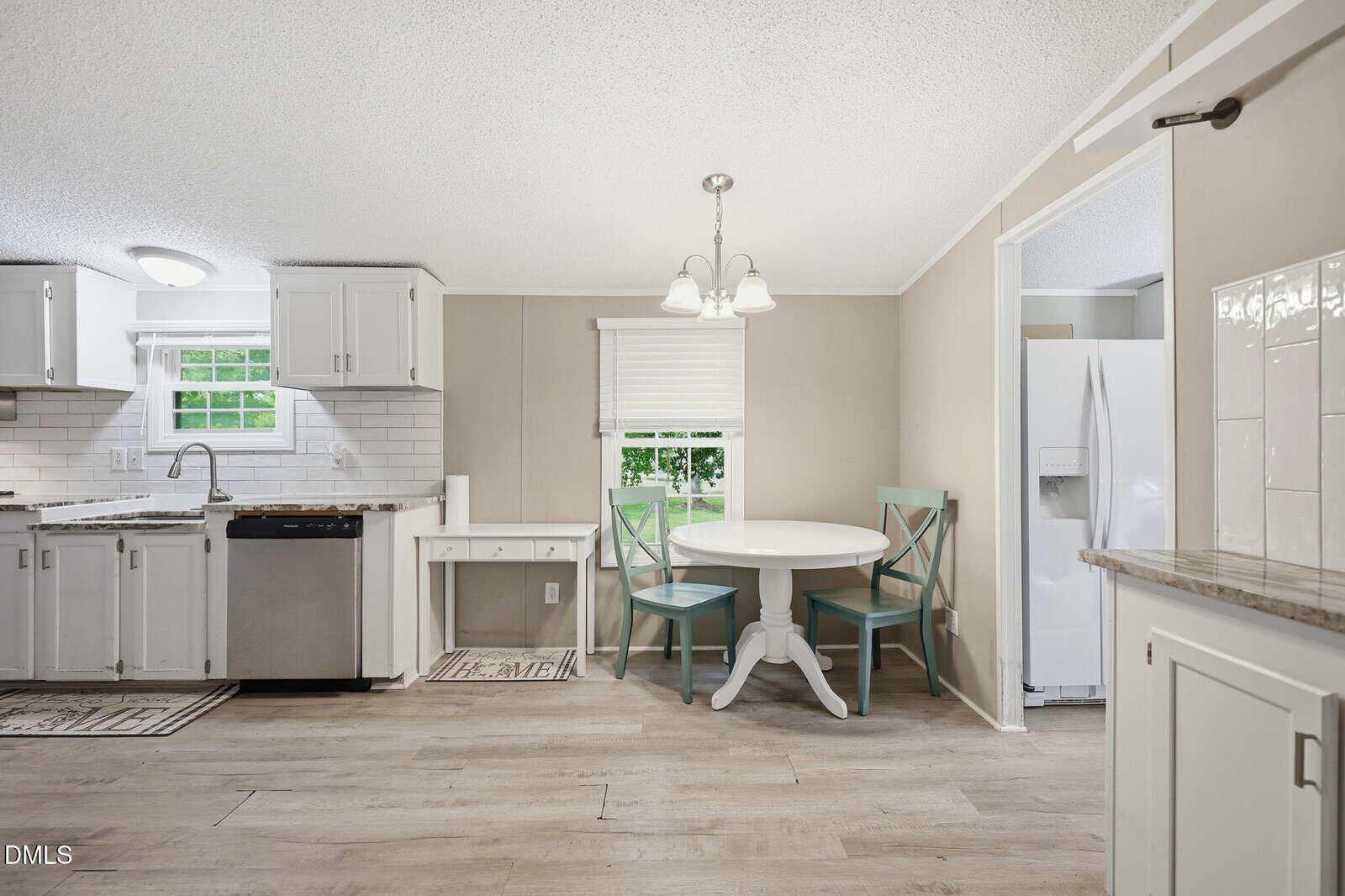 Bright kitchen with white cabinets, stainless steel appliances, a round dining table with two chairs, and a window with a shade. Light wood flooring and neutral walls create an inviting, modern atmosphere.