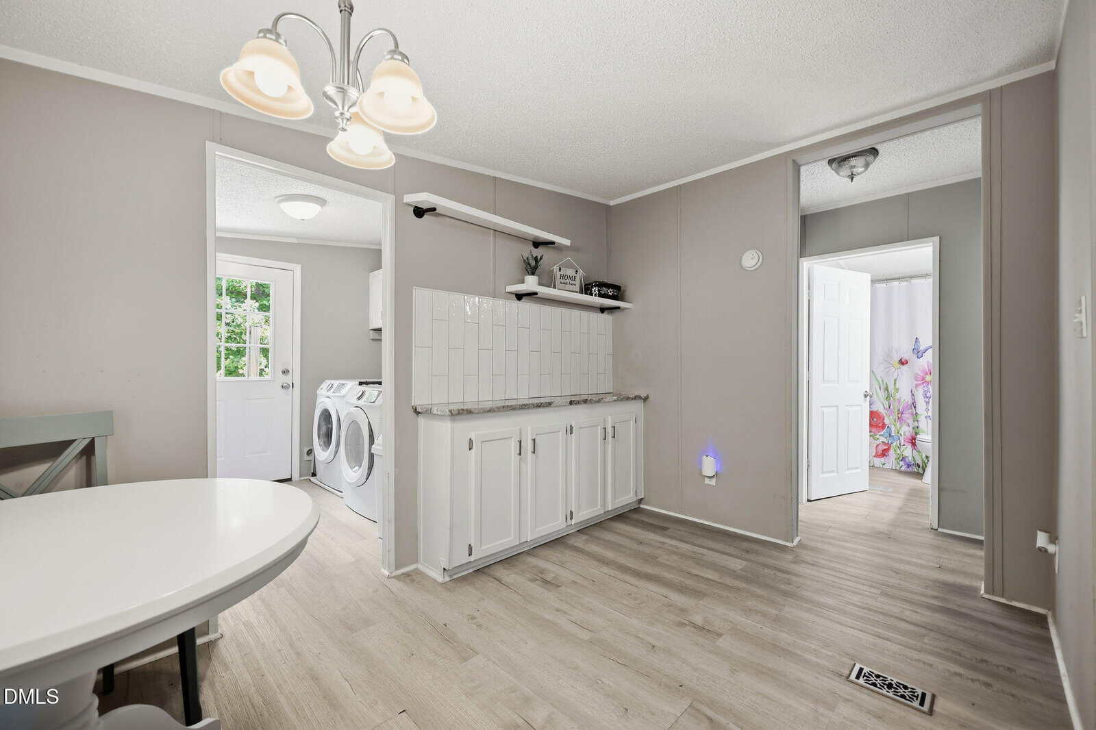 Modern kitchen with light wood floors, white cabinetry, a small dining table, washing machine and dryer in the adjacent laundry room, and a bathroom with a floral shower curtain visible through an open door.