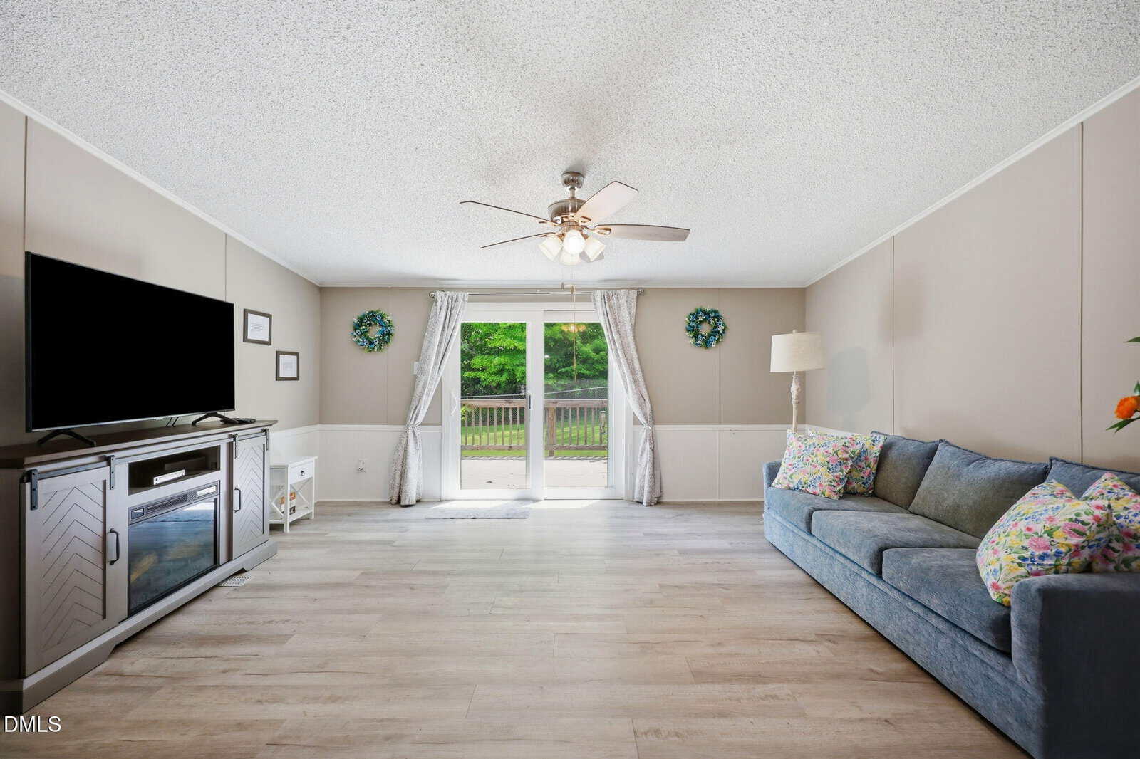 Bright living room with light wood flooring, a blue couch, a TV on a console, a standing lamp, and double glass doors leading to a deck with green trees outside. Neutral walls and decorative accents provide a cozy feel.