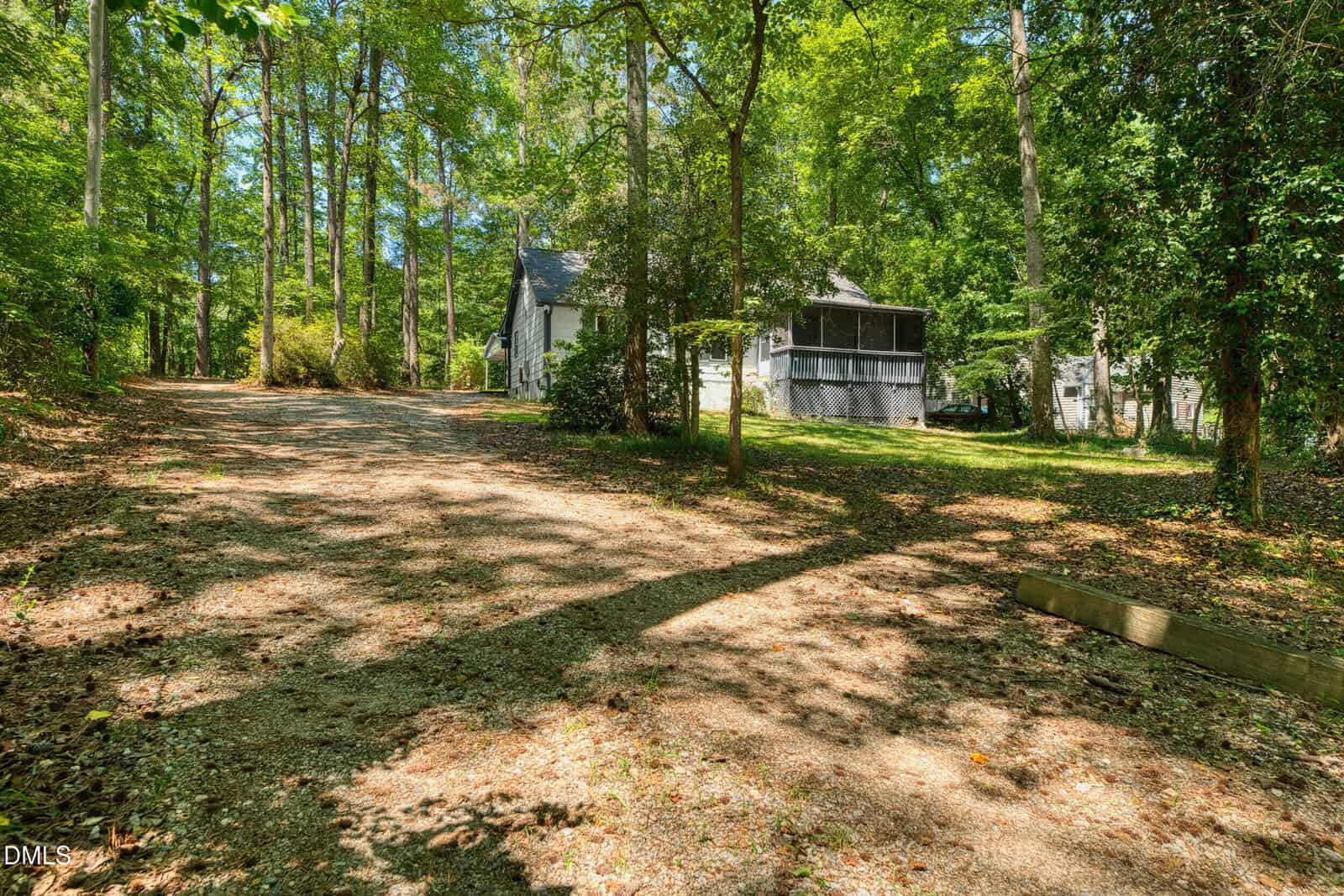 A gravel driveway leads through a wooded area to a white house with a screened porch, surrounded by tall trees and greenery on a sunny day.