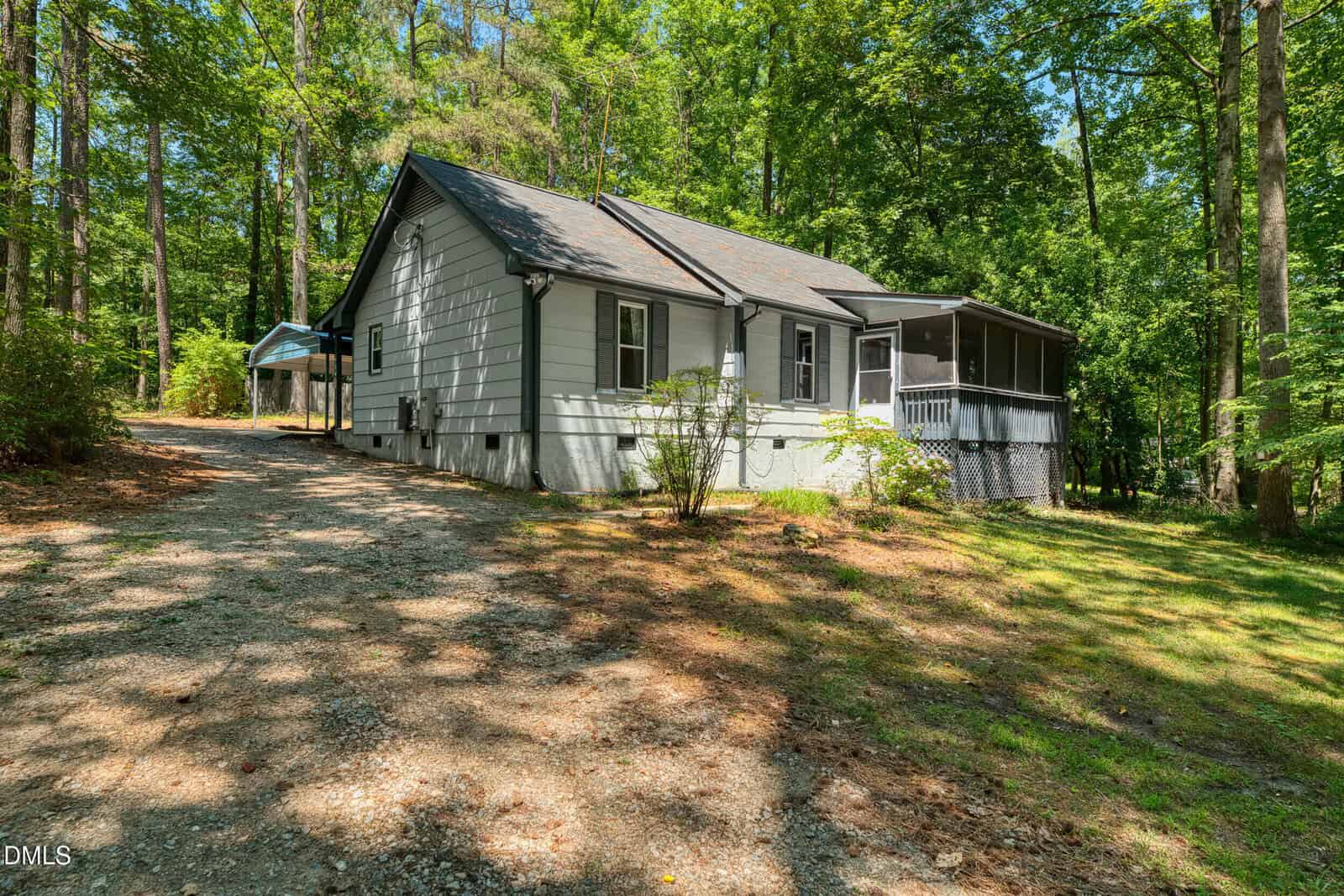 A small white house with a screened porch sits among tall trees on 956 Holden Road in Youngsville, surrounded by greenery. A gravel driveway leads up to the house, which is partially shaded by sunlight filtering through the trees.