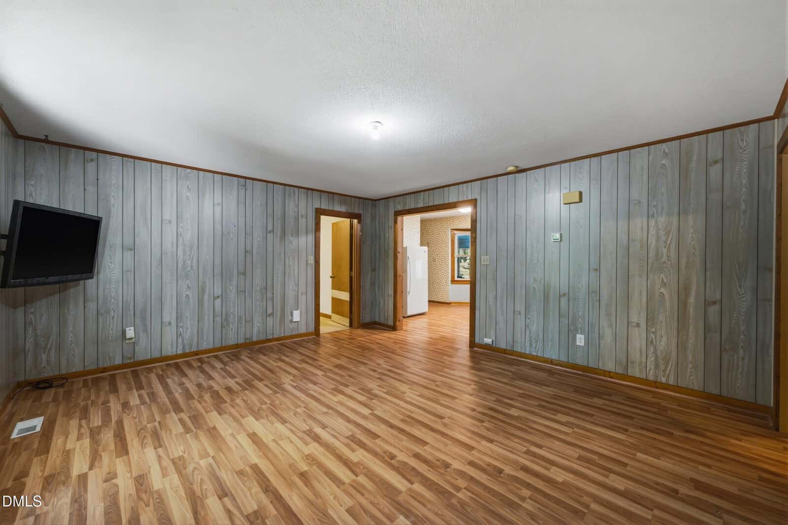 Empty room with wood-paneled walls, laminate wood flooring, a wall-mounted TV, and open doorways leading to a kitchen with white appliances and another adjacent room. Ceiling light fixture is centered.