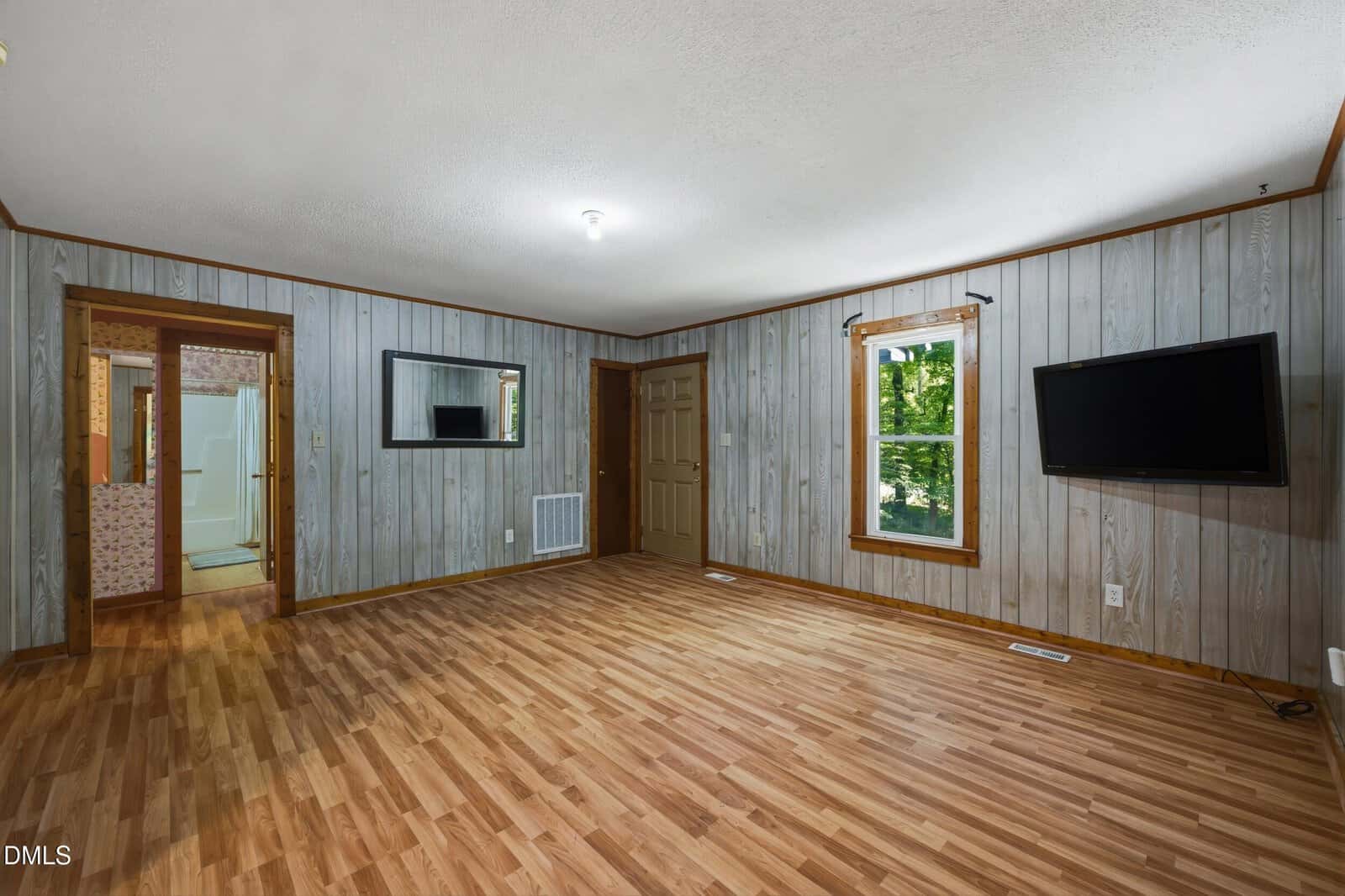 A living room with wood panel walls, a wood floor, a window, a wall-mounted TV, a mirror, and a door. An open doorway shows a glimpse of a bathroom and another room.