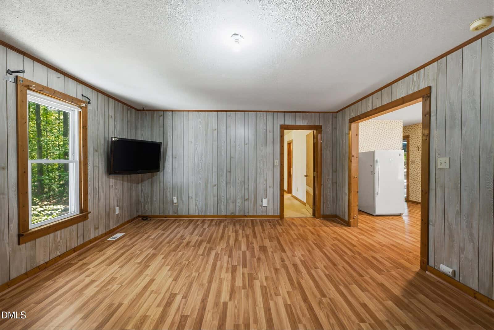 A room with wood-paneled walls and wood-style flooring, featuring a wall-mounted TV, two windows, and open doorways leading to a hallway and a kitchen with a white refrigerator.