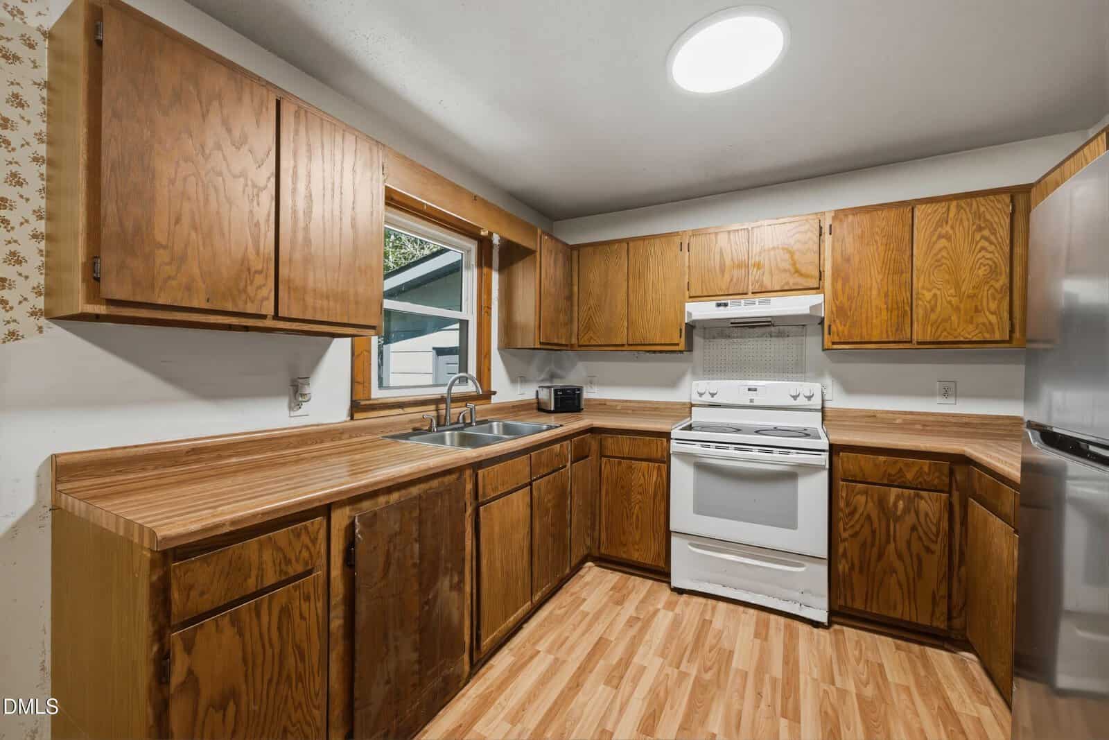 Kitchen with wooden cabinets and countertops, white stove and oven, stainless steel sink, refrigerator, wood-patterned floor, window above sink, and a round ceiling light.