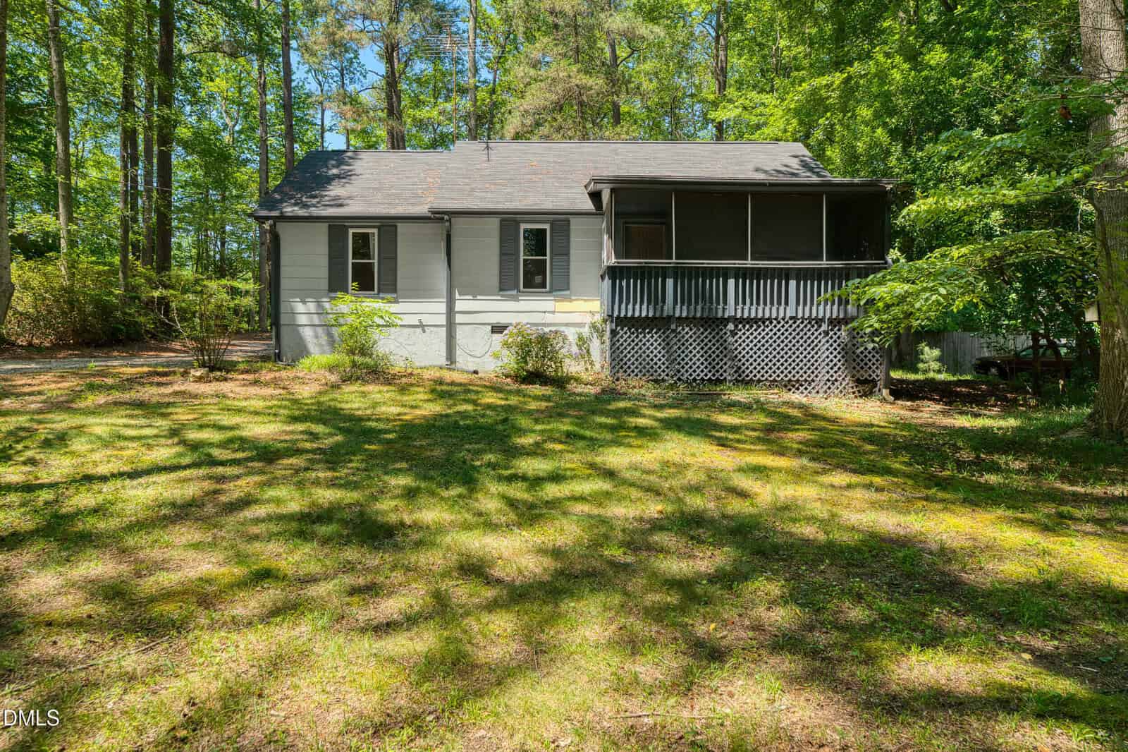 A small gray house with a screened porch, surrounded by trees and greenery, sits in a sunlit yard with grass and patches of shade.