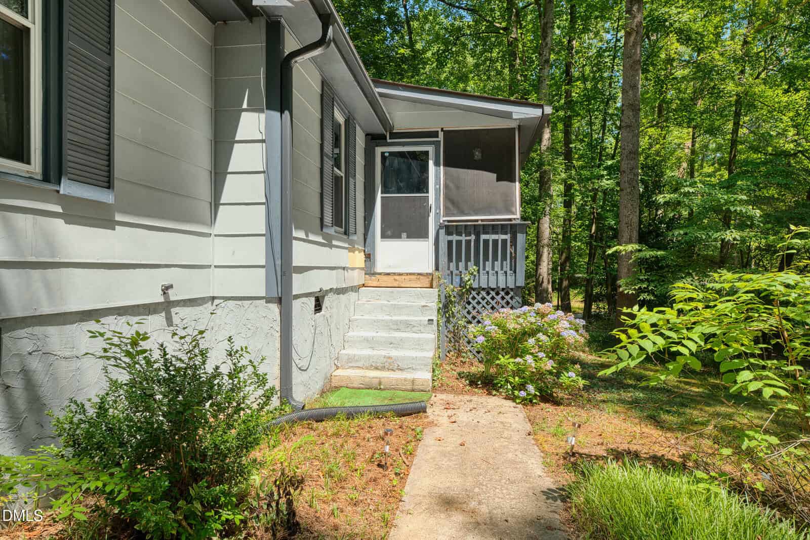 A concrete path leads to a set of stairs up to a white house with gray shutters, a screened porch, and surrounded by lush green trees and shrubs on a sunny day.