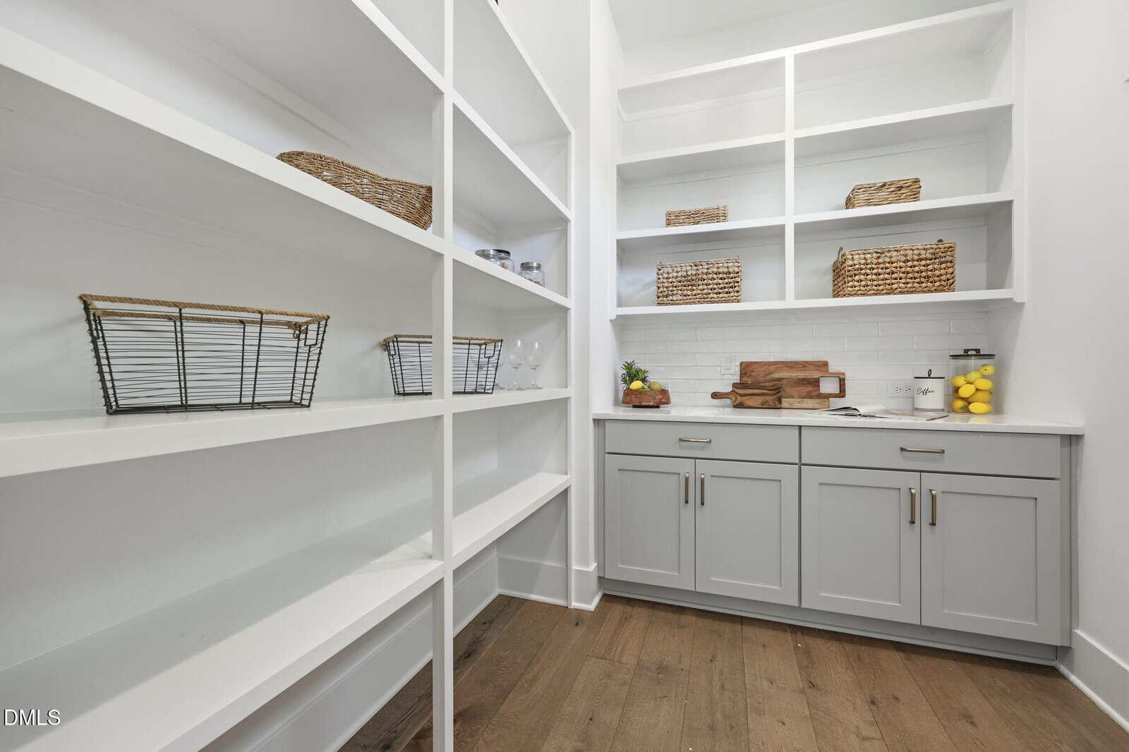 A walk-in pantry with white shelves, gray lower cabinets, wire and wicker baskets, wooden cutting boards, and a glass jar of lemons on a hardwood floor. The space is bright and organized.