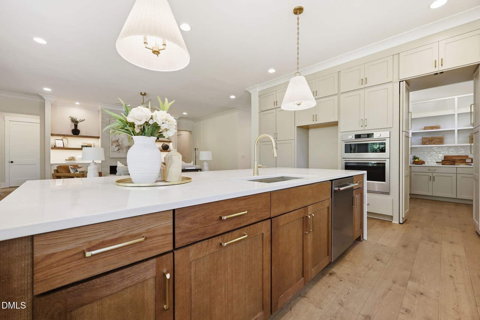Modern kitchen with a large island featuring a sink, wood cabinets with gold handles, and white countertops. Pendant lights hang above. In the background are beige cabinets, double ovens, and an open pantry. A vase with flowers decorates the island.